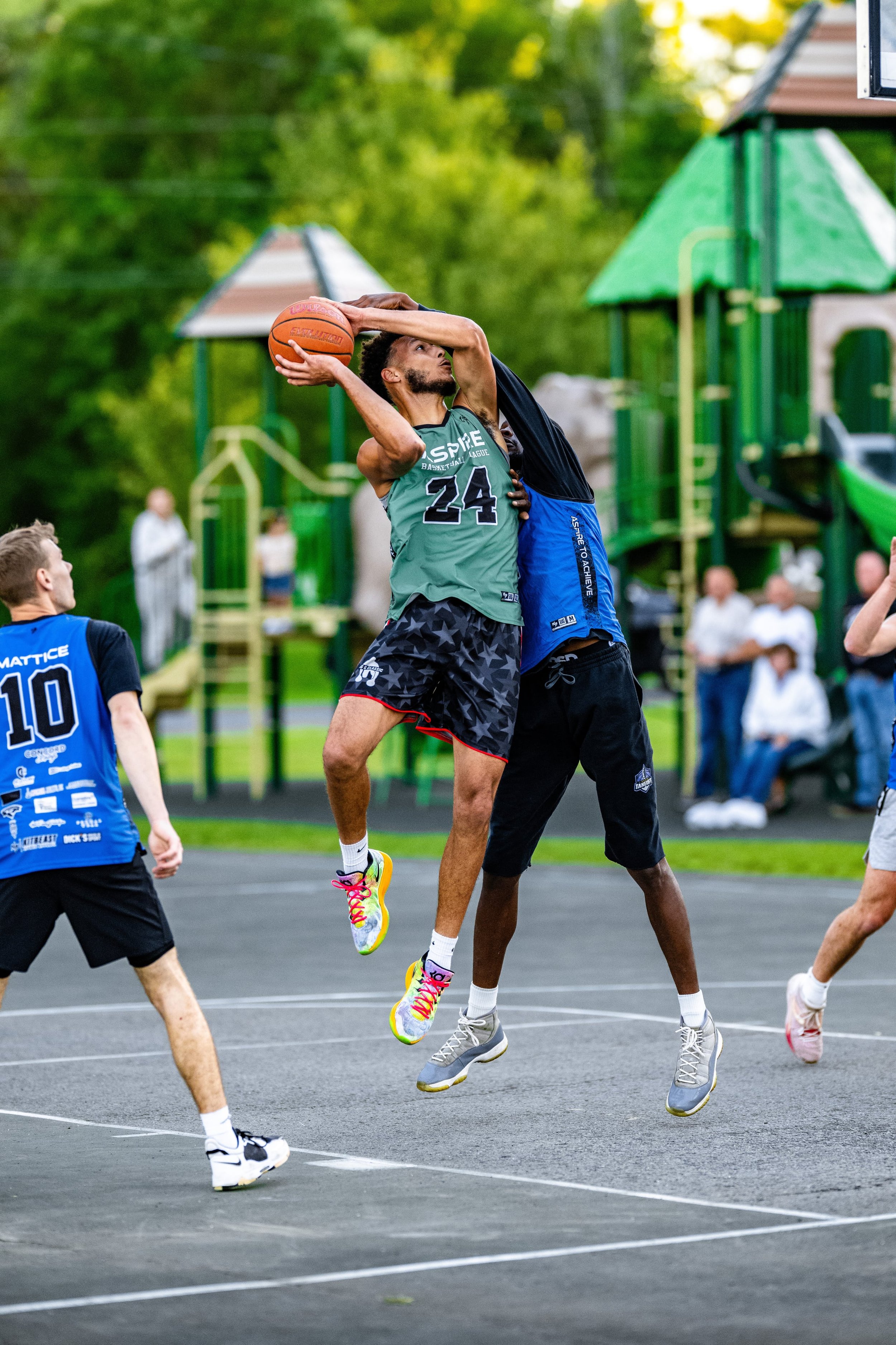 Basketball game with players jumping for ball on outdoor court, playground and spectators in background.