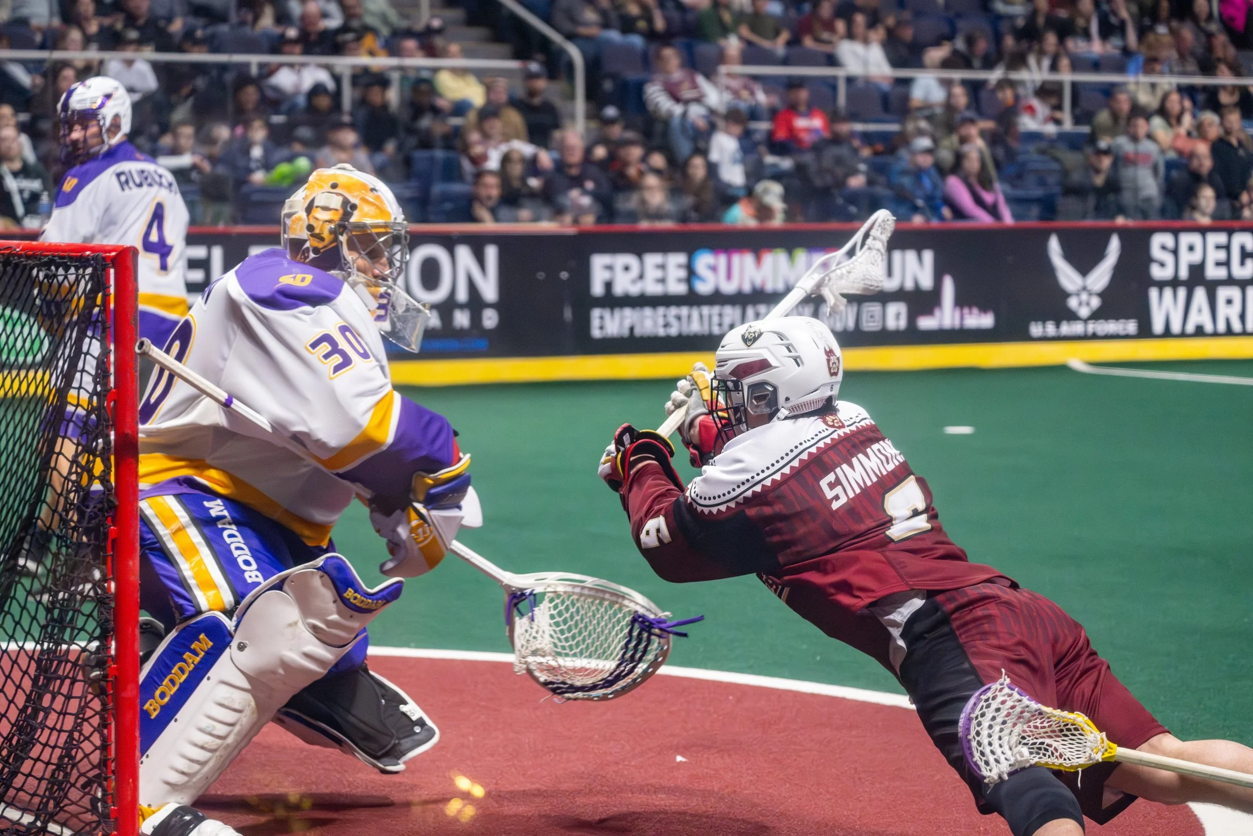 Lacrosse game with goalkeeper attempting to block a shot from a player in maroon uniform