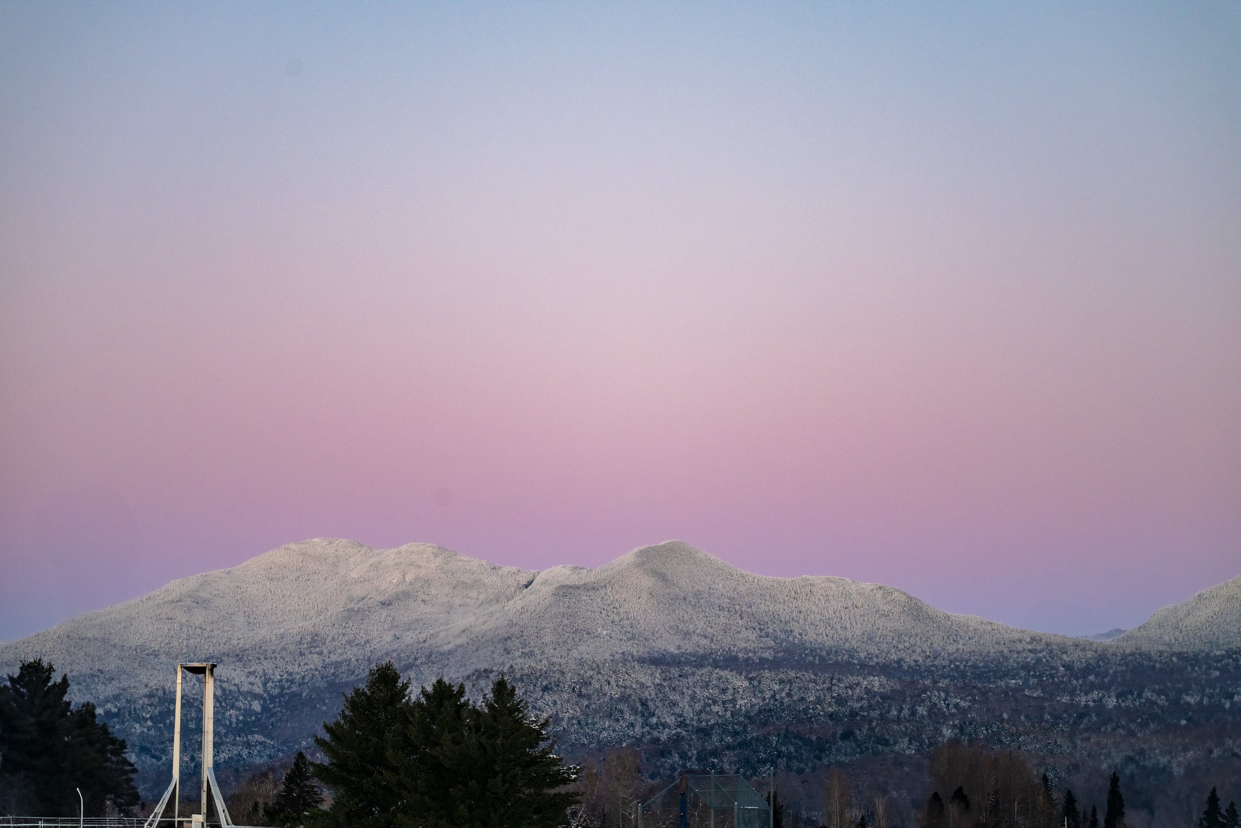 Snow-covered mountain range at sunset with a pink and purple sky, trees in the foreground, and a tennis court structure visible at the bottom left.
