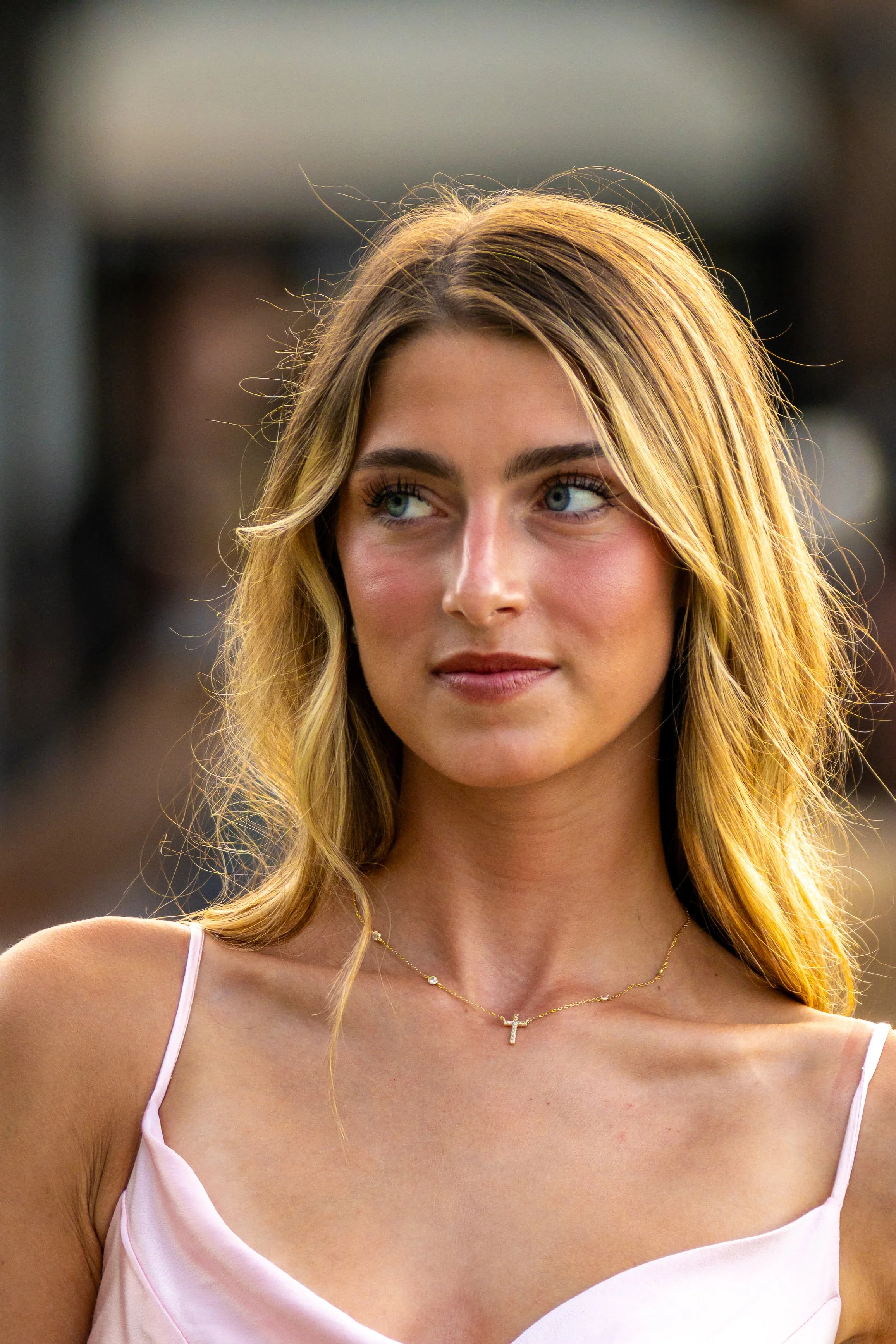 A young woman with long blonde hair and blue eyes, wearing a pink spaghetti strap top and a delicate gold necklace with a cross pendant, standing outdoors.