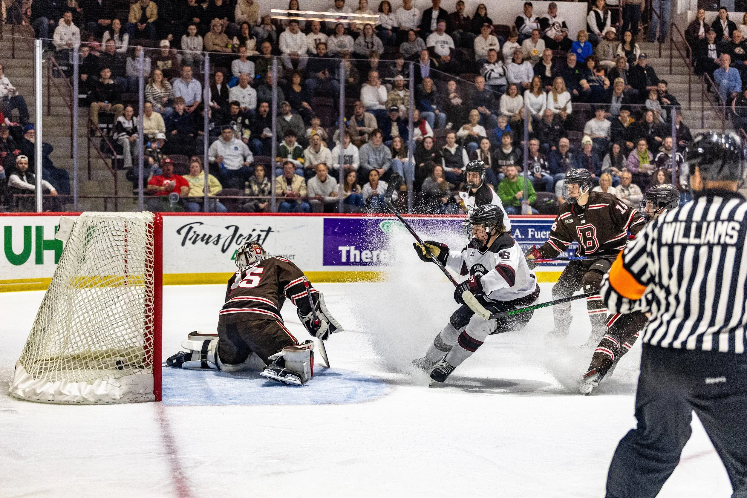 An ice hockey game with players in action near the goal, with a goalie in black kneeling and preparing to block the puck, spectators watching from the stands, and a referee in black and white stripes in the foreground.