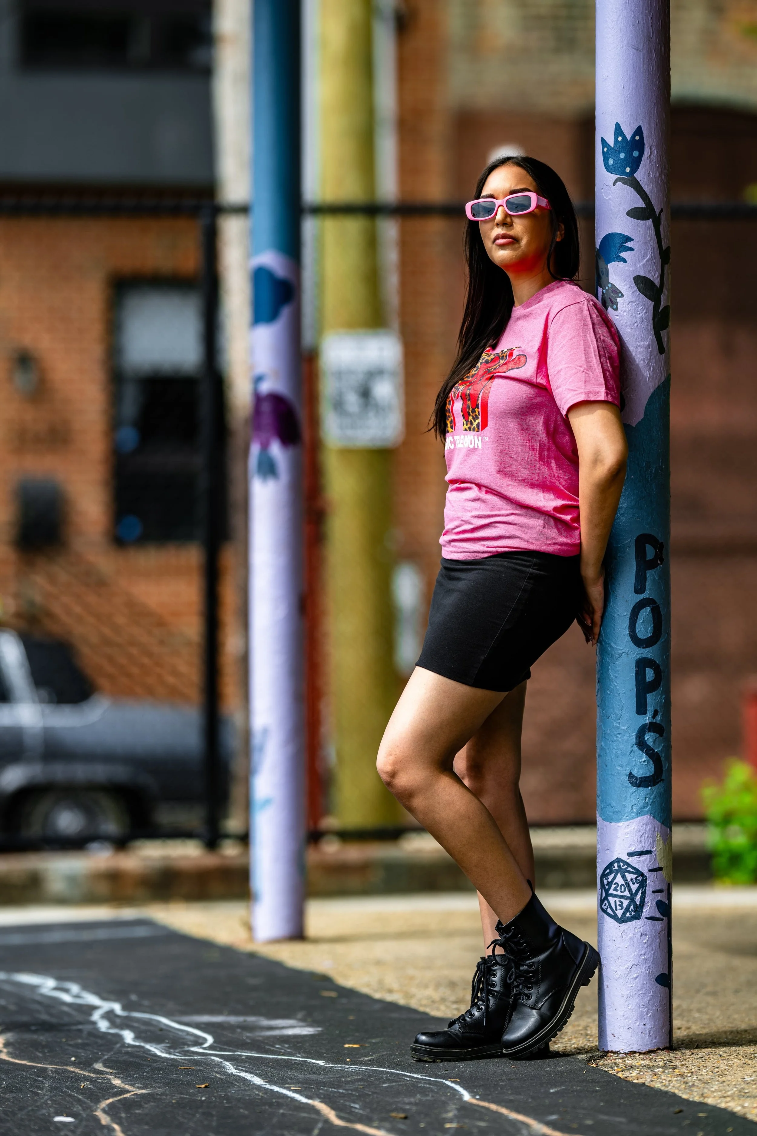 A woman with long dark hair wearing pink sunglasses, a pink graphic t-shirt, black shorts, and black combat boots leaning against a blue pole with colorful painted designs on an outdoor basketball court.