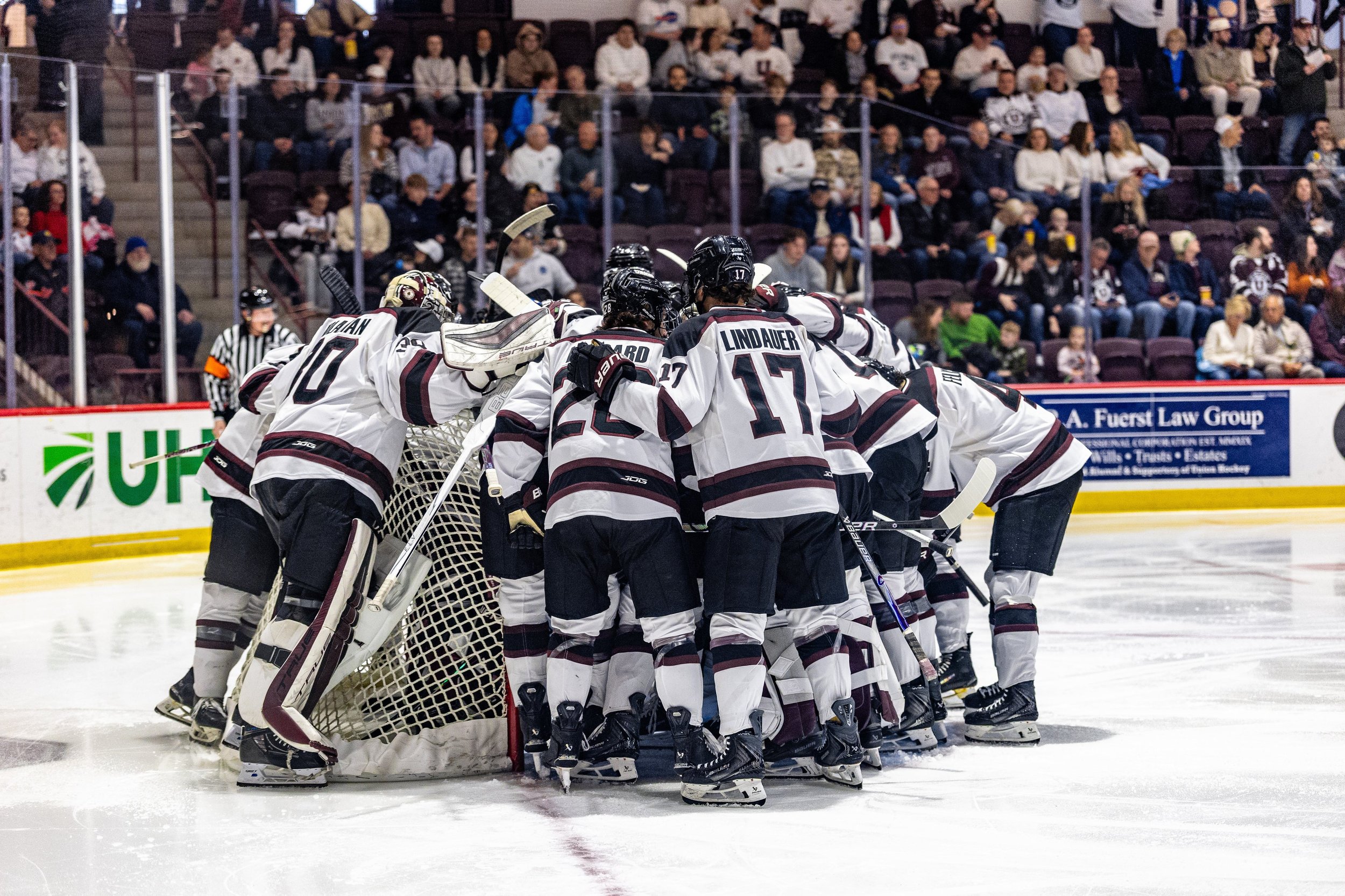 Hockey team in white jerseys huddles together near the goal post on ice rink while spectators watch from stands.