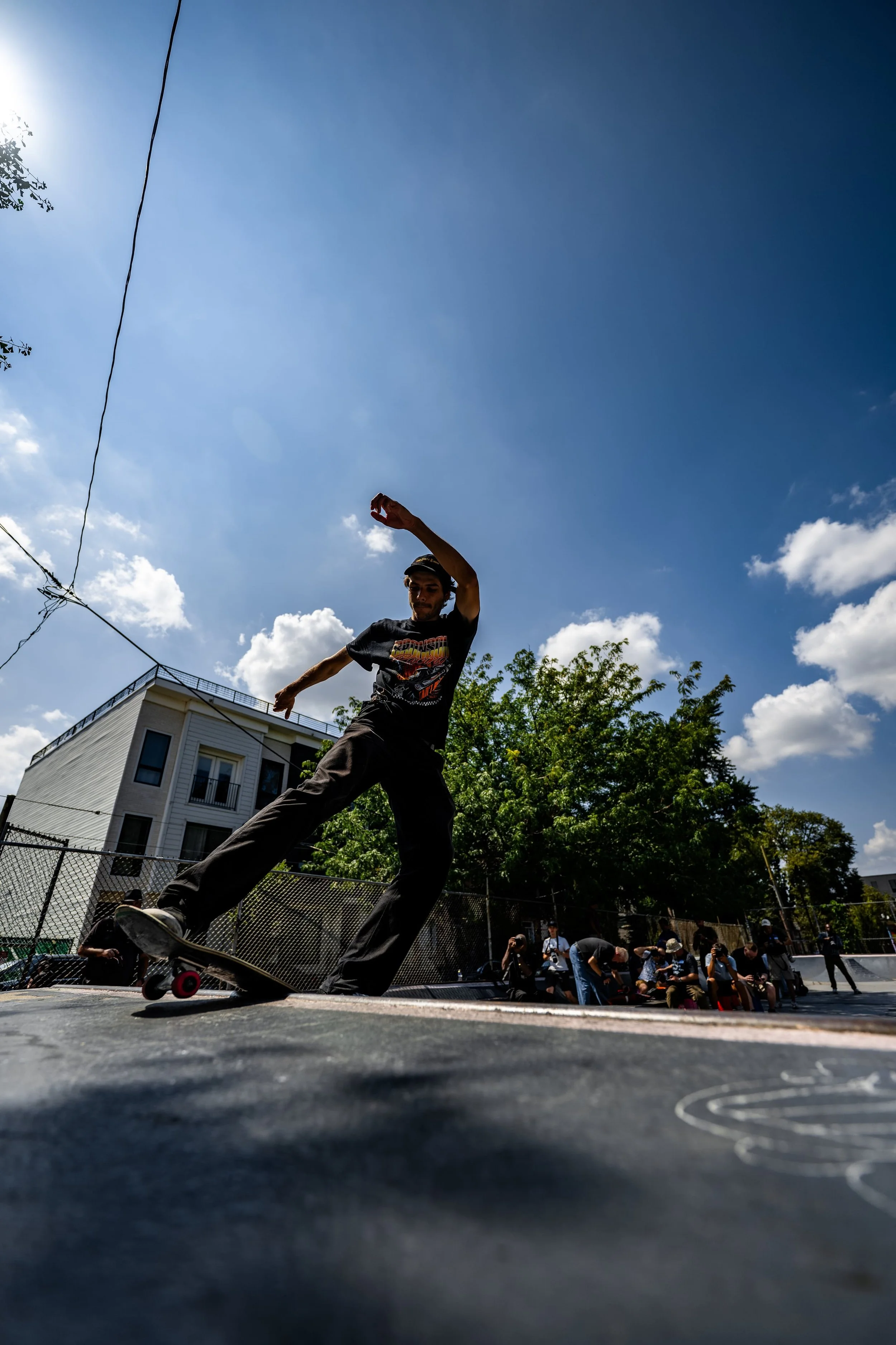 Skateboarder performing a trick on a skateboard at an outdoor skate park with a group of people sitting along the fence, trees, a building, and a blue sky with clouds in the background.