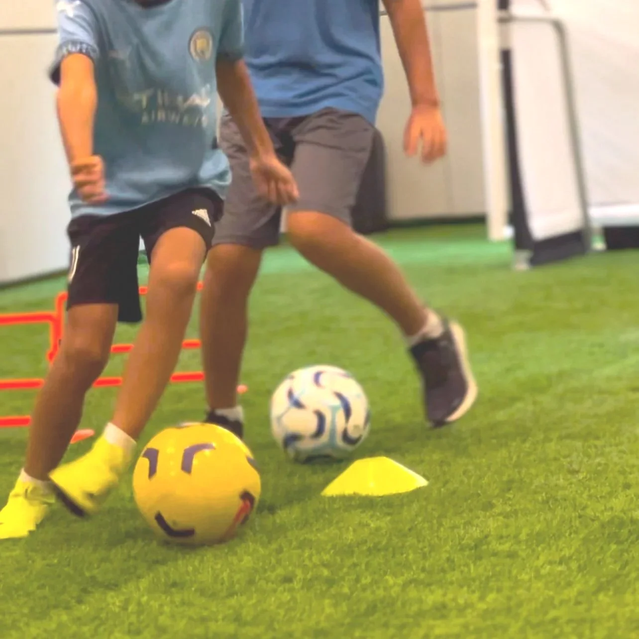 Two boys practicing soccer drills with cones and balls on an indoor turf field.
