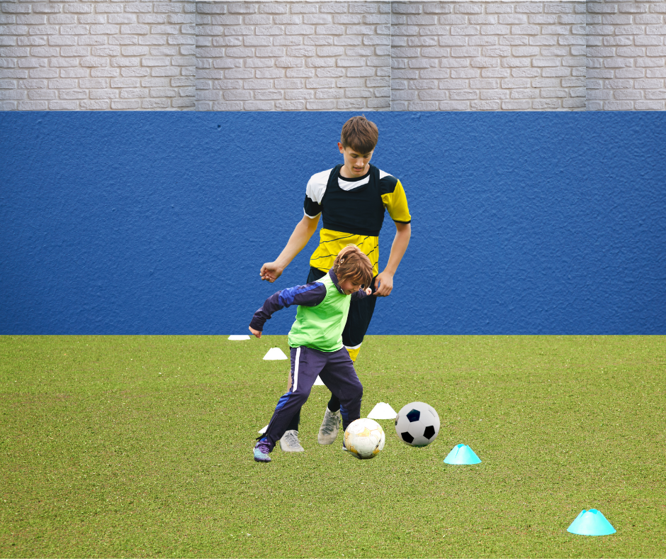 A young boy practicing soccer drills with a coach on a grassy indoor turf, using soccer balls and training cones.