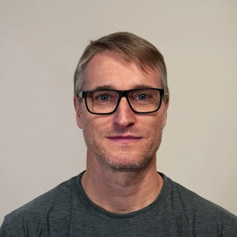 Adam Thomas. A man with light skin, short light brown hair, wearing black-rimmed glasses and a dark gray t-shirt, smiling slightly against a plain beige background.