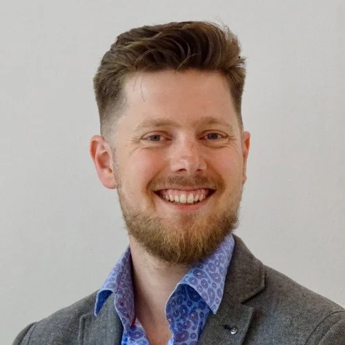 Joris de Vries. A man with short, light brown hair and a beard, smiling, wearing a blue dress shirt and a dark gray blazer.
