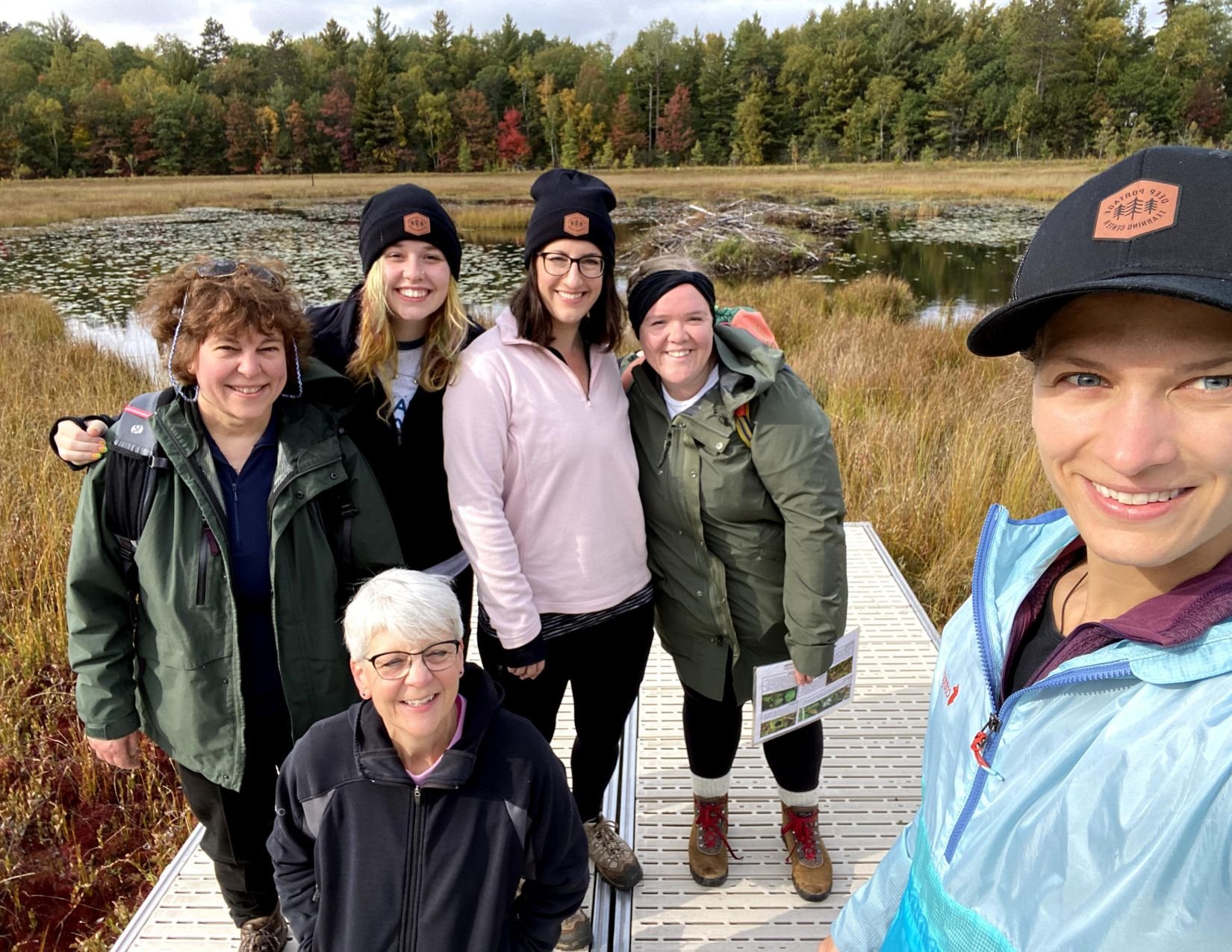Group of six women on a bog boardwalk with autumn-colored trees in the background.