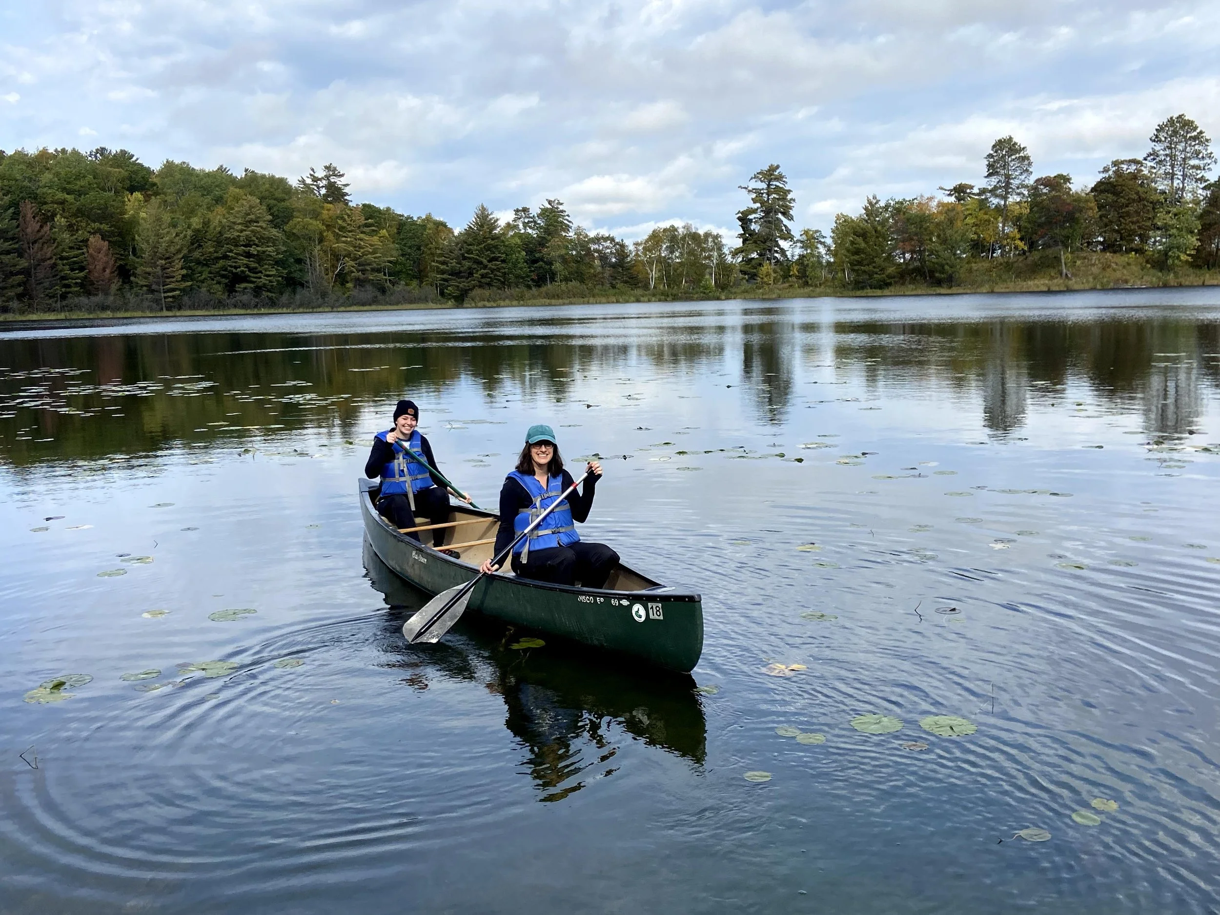 Two people canoeing on a calm lake with trees and cloudy sky in the background.