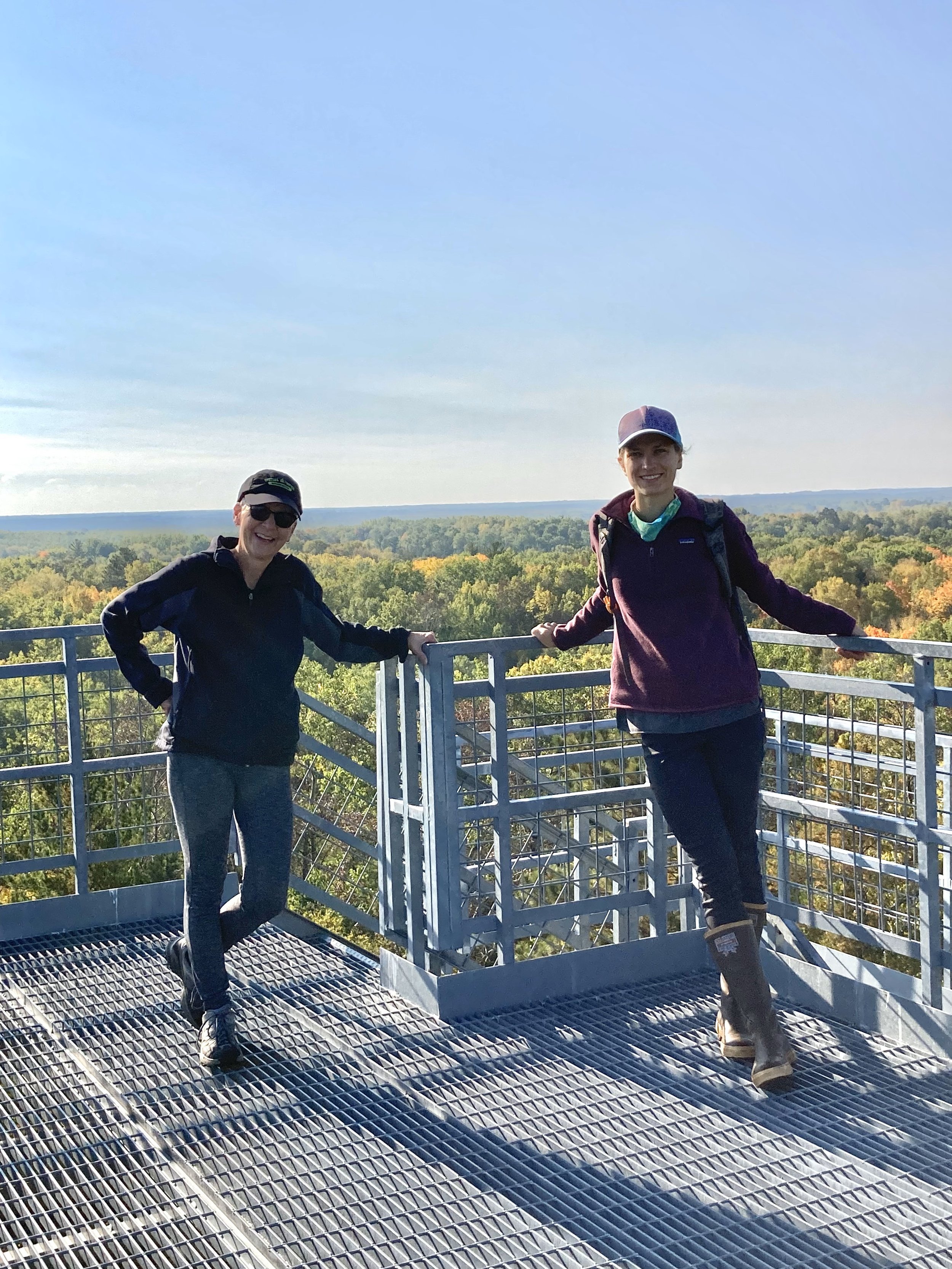 Two smiling women standing on a metal observation deck with a scenic view of trees and a clear sky in the background.