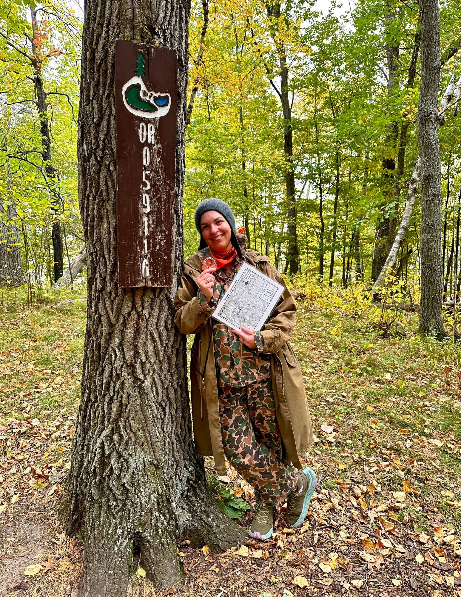 A woman in camouflage clothing and a brown jacket standing in a wooded area, smiling, holding a map and a small item, leaning against a tree with a brown marker that reads 'OR 59116'