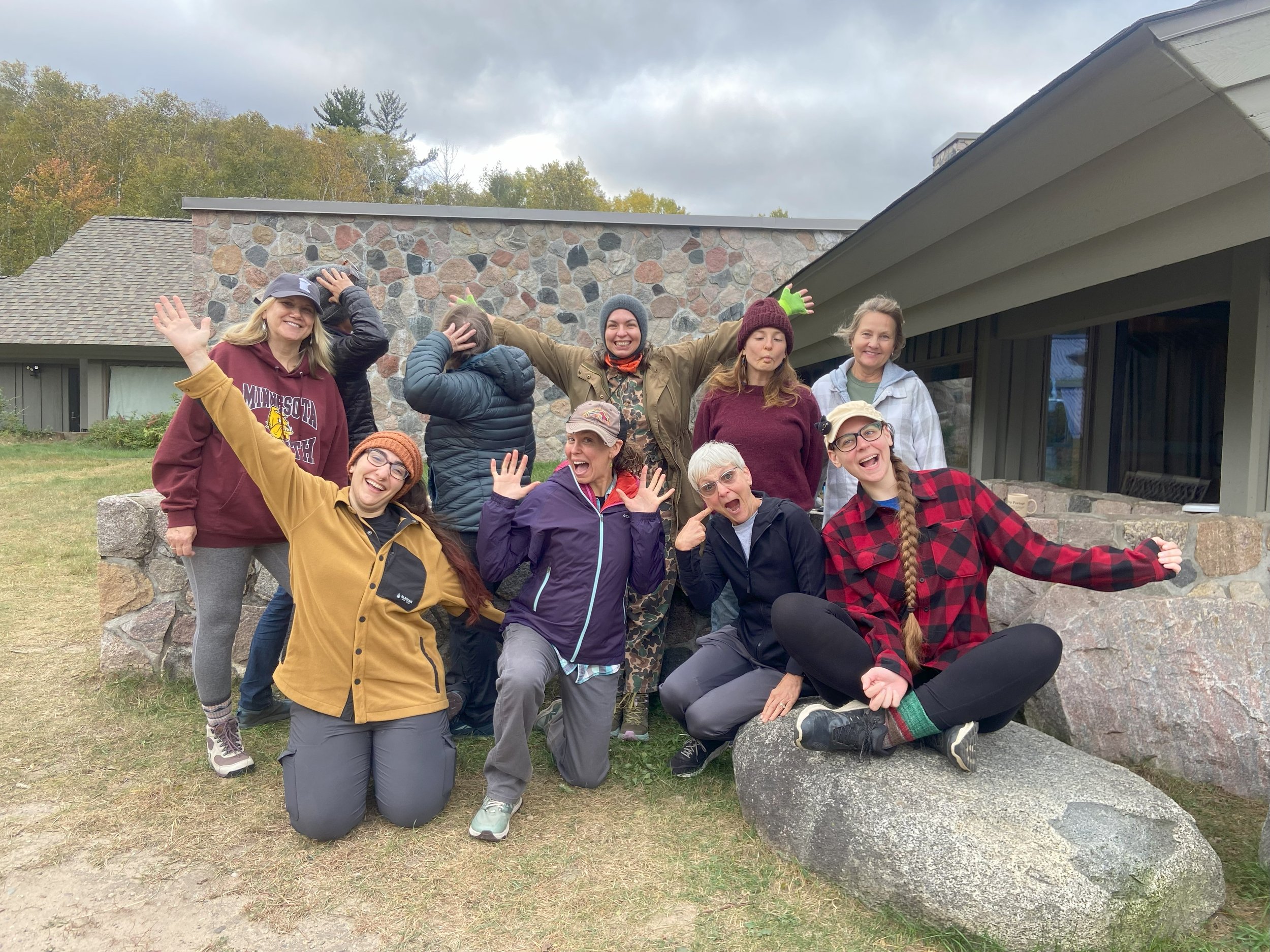 A group of ten women posing outdoors in front of a stone and wooden building, some smiling, some making silly gestures, wearing casual outdoor clothing, with trees and cloudy sky in the background.