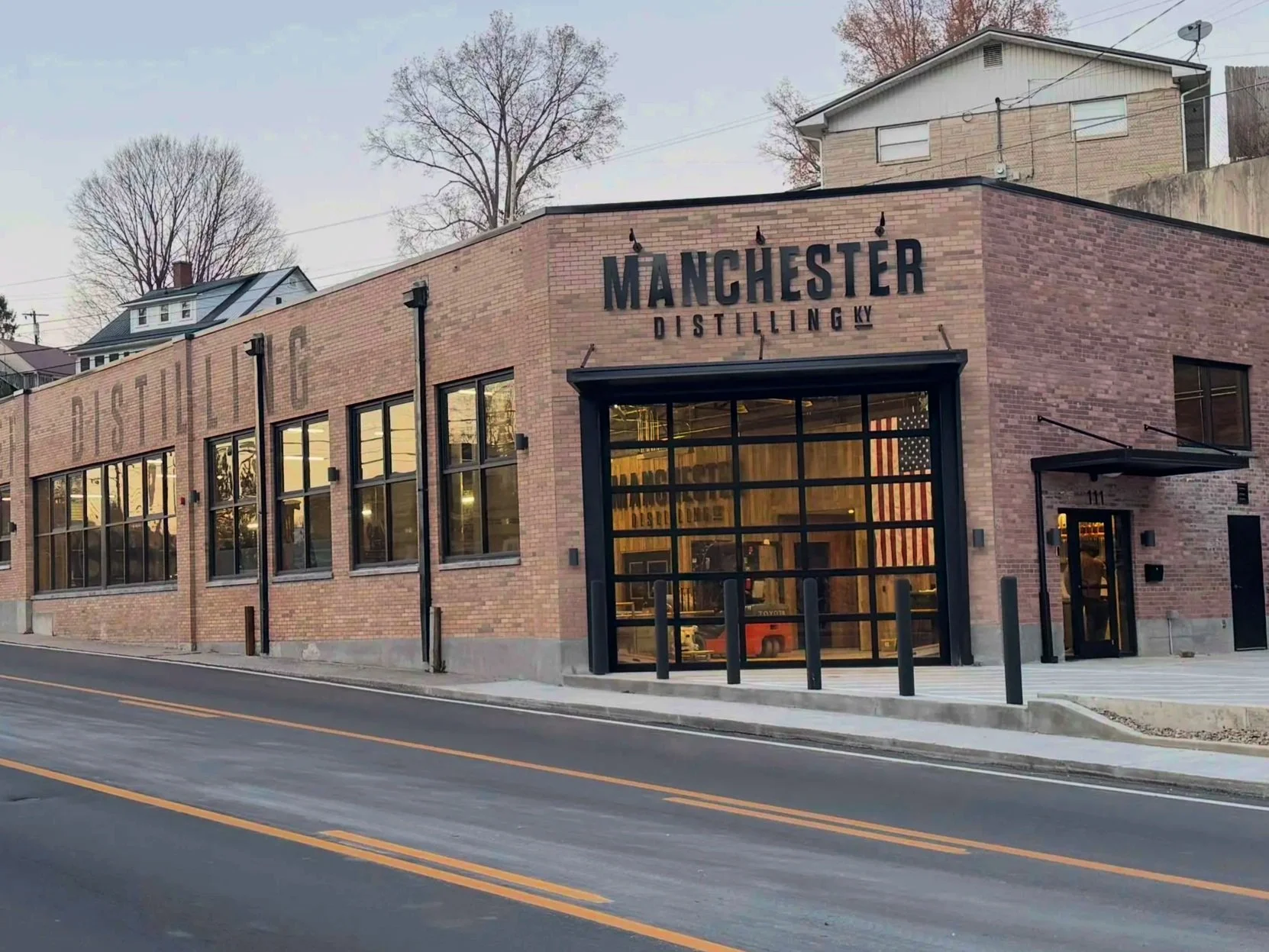 Exterior view of a brick building with large windows, labeled 'Manchester Distilling KY,' on a street at sunset.