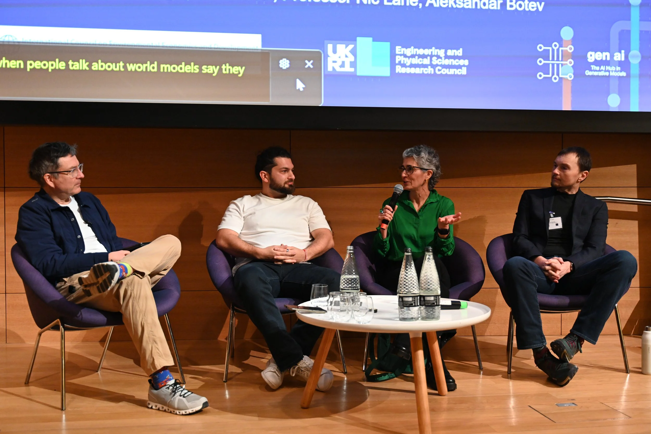 Four people seated in front of a screen and behind a coffee table with water bottles on it. One is holding a microphone