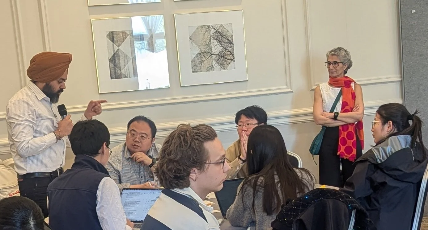 Person with a microphone standing with other people sitting round a table