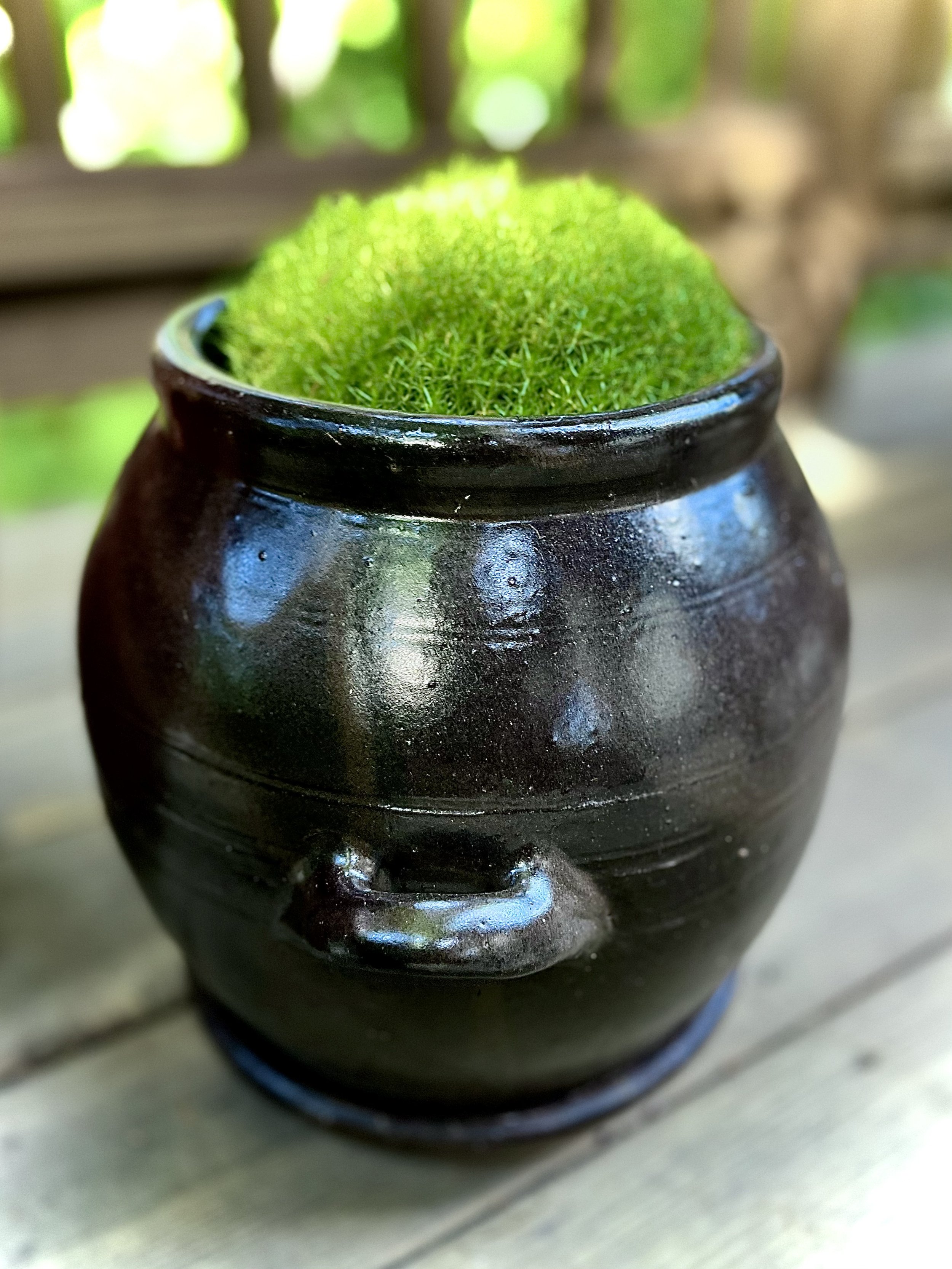 A small dark ceramic pot with a face-like design, filled with bright green moss, placed on a wooden surface outdoors.