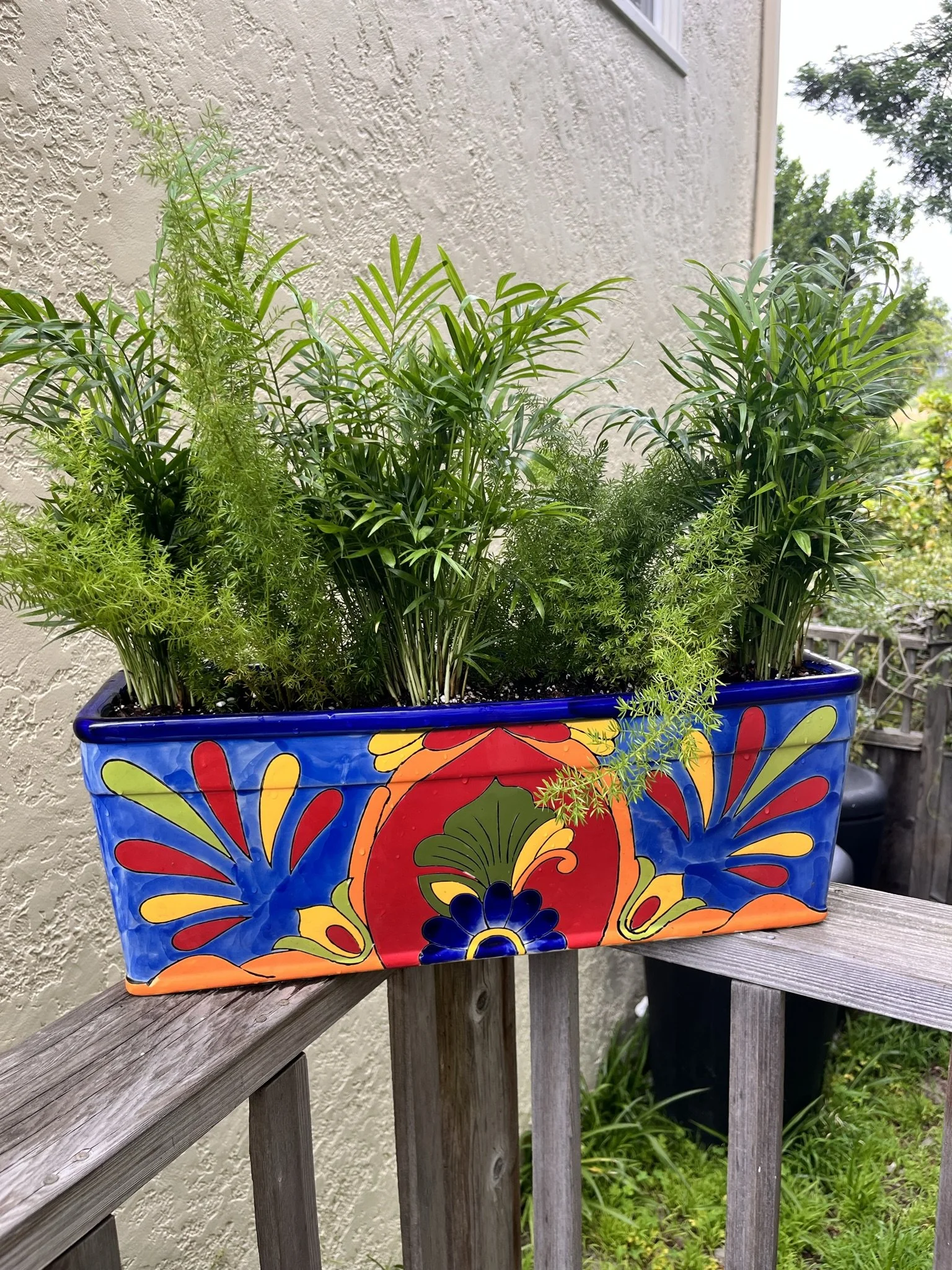 A rectangular, colorful ceramic planter filled with green herbs, sitting on a wooden railing outside near a beige wall and some trees in the background.