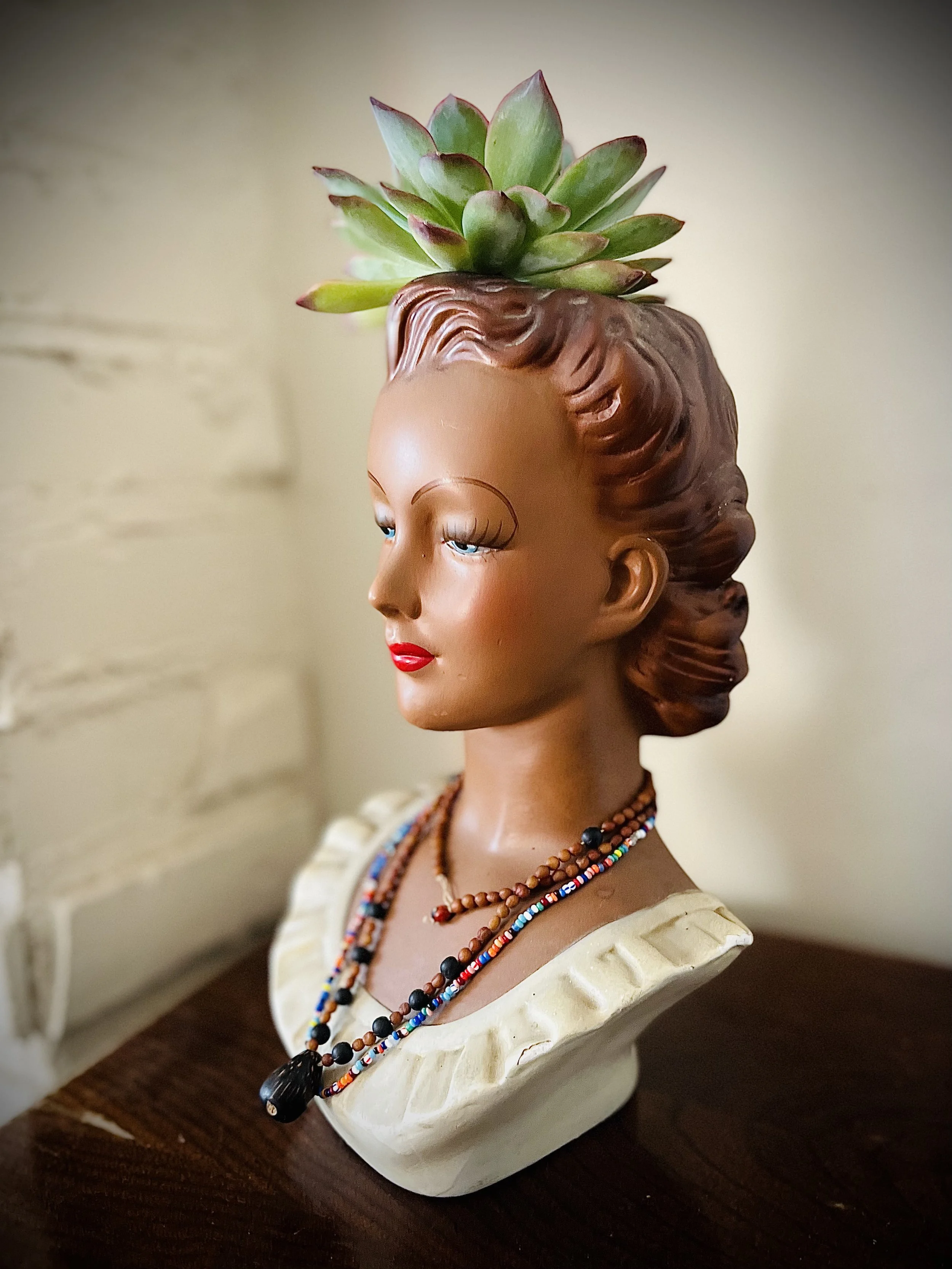 A vintage bust of a woman with a succulent plant on her head, wearing layered beaded necklaces, and dressed in a cream-colored top, placed on a dark wooden surface, with a light-colored wall in the background.