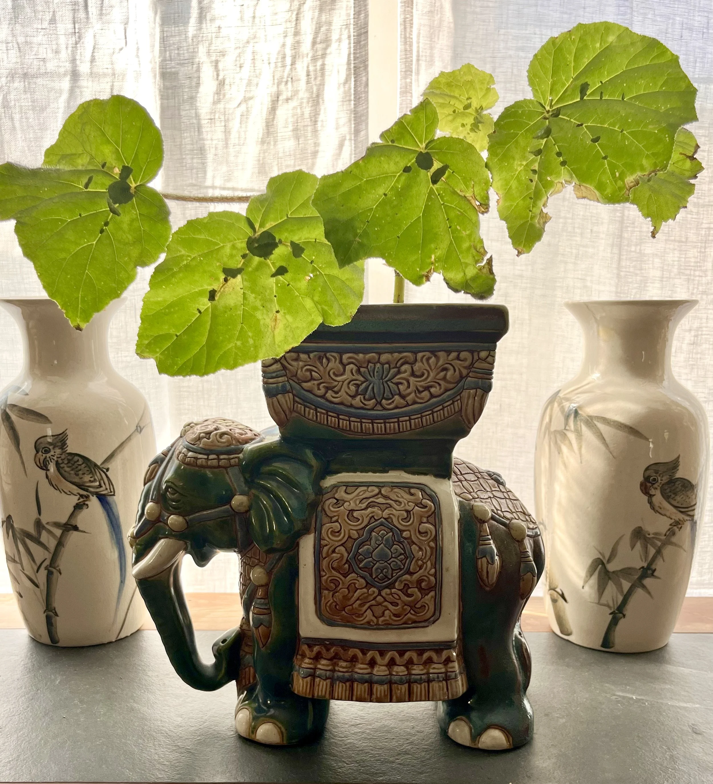 Decorative ceramic elephant-shaped planter with green leaves, flanked by two white vases with bird and bamboo designs, set against a sheer curtain background.