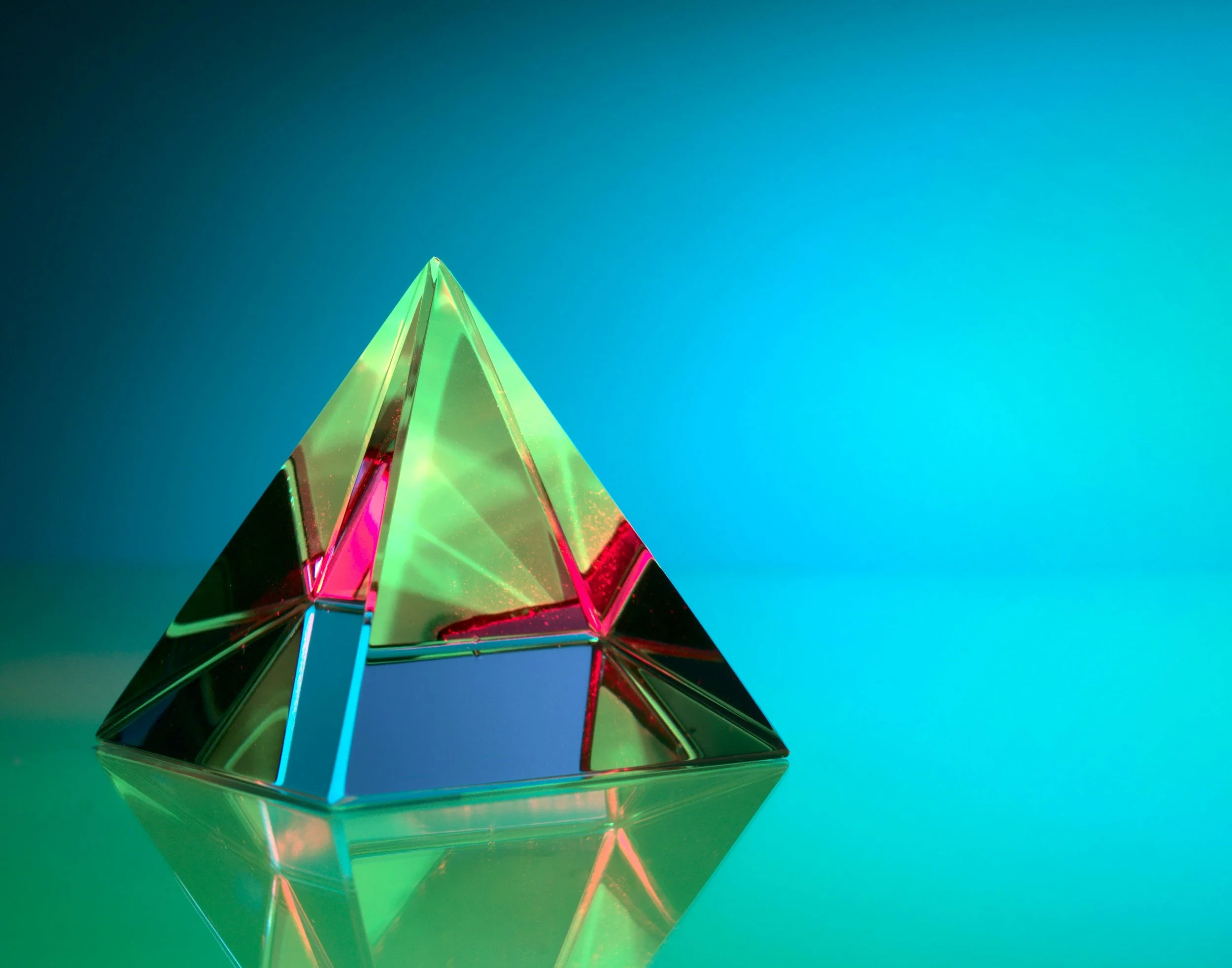 A colorful glass pyramid sculpture reflecting light, placed on a reflective surface against a gradient blue-green background.