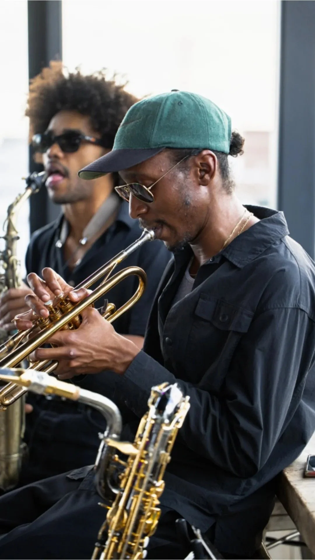 Two musicians playing saxophones, one in the foreground wearing glasses and a green hat, and the other in the background with sunglasses and curly hair, both dressed in dark clothing, inside Lucky Buck's brightly lit rooftop bar. 