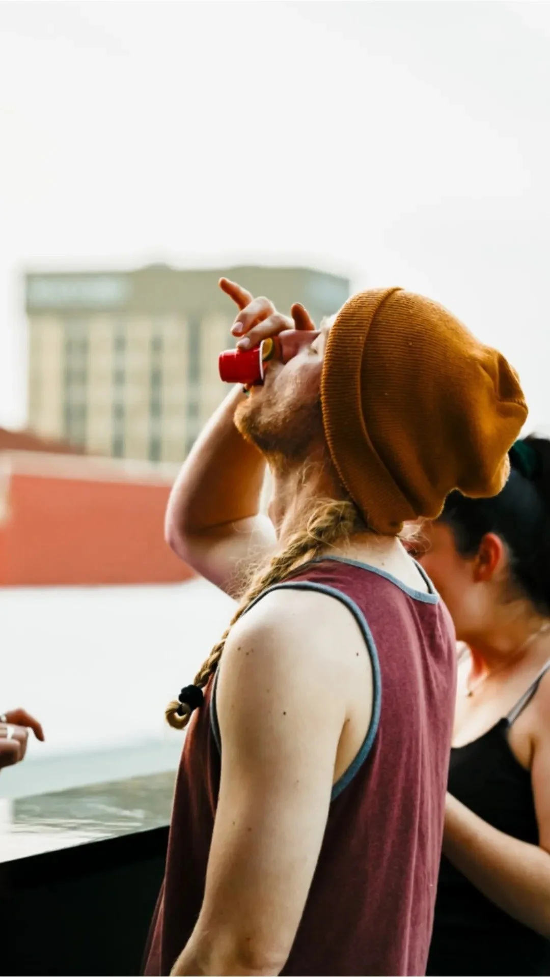 A young man with long red hair, a beard, and a brown beanie, wearing a sleeveless maroon shirt with grey trim, is drinking from a small red cup. He has a braided hairstyle at Luck Buck's Rooftop Bar with tall buildings in the background, along with a