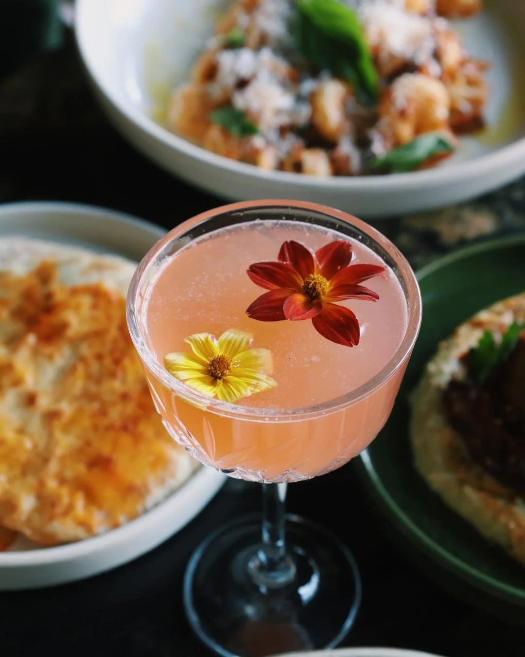 A pink cocktail with two edible flowers floating in the top, several dishes surround it on a black marble counter top
