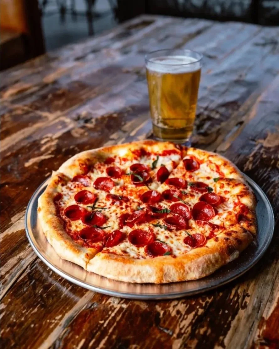 A pepperoni pizza on a metal tray with a glass of beer on a rustic wooden table.