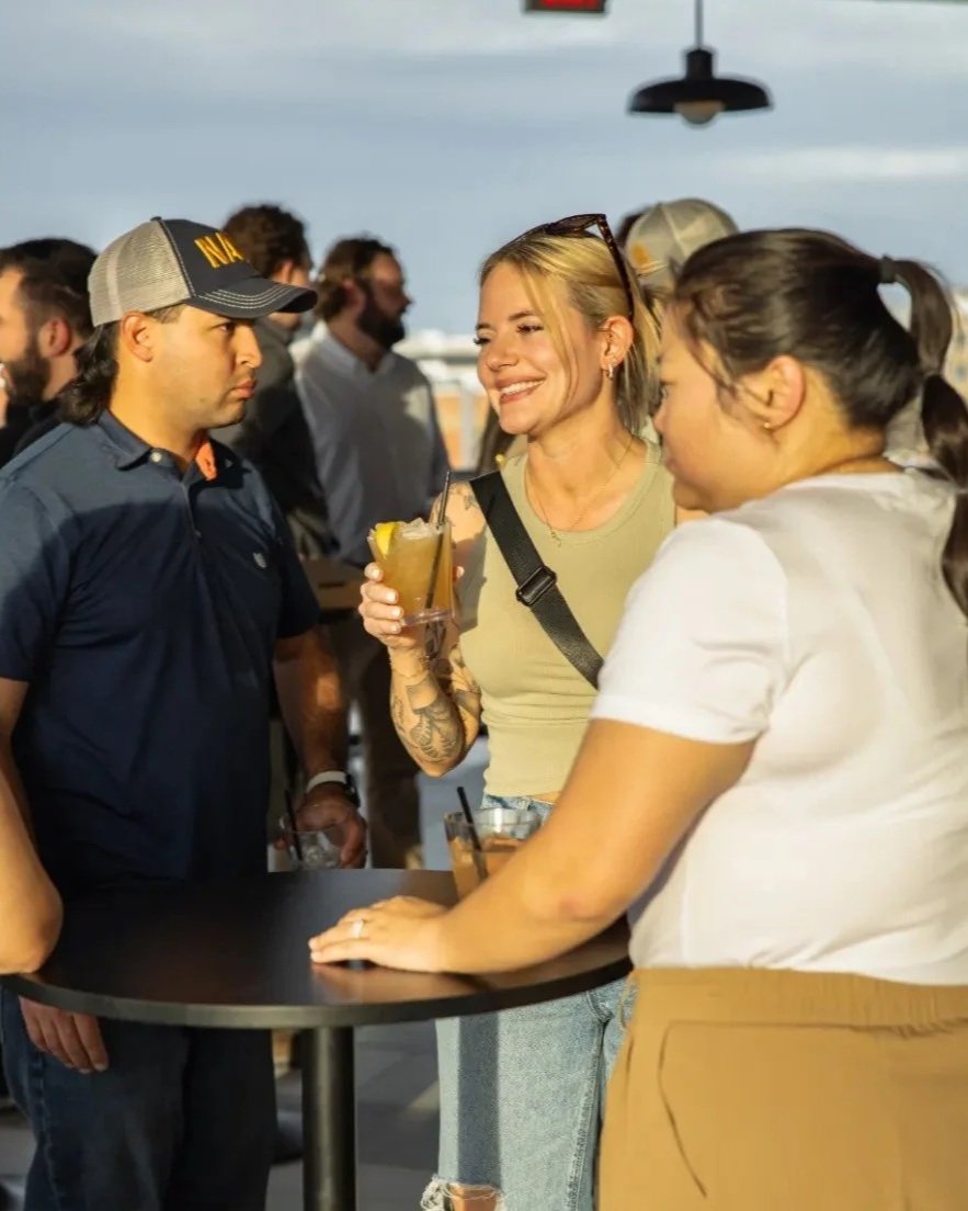 People socializing at Lucky Buck's Rooftop Bar, with a woman smiling holding a drink, and two women and a man around a high table, in the evening with cityscape in the background.