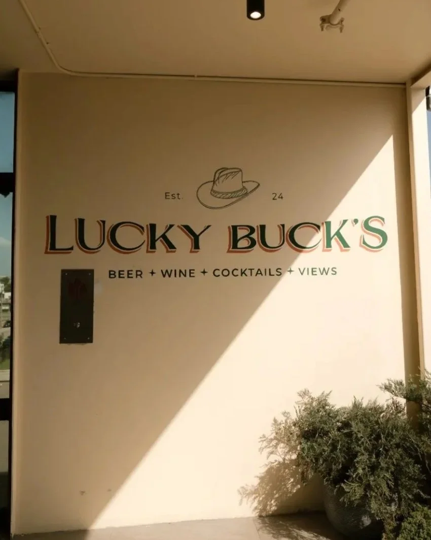 Sign for Lucky Buck's bar featuring a hat drawing, established in 2024, offering beer, wine, cocktails, and views, with plants and sunlight shadows nearby.