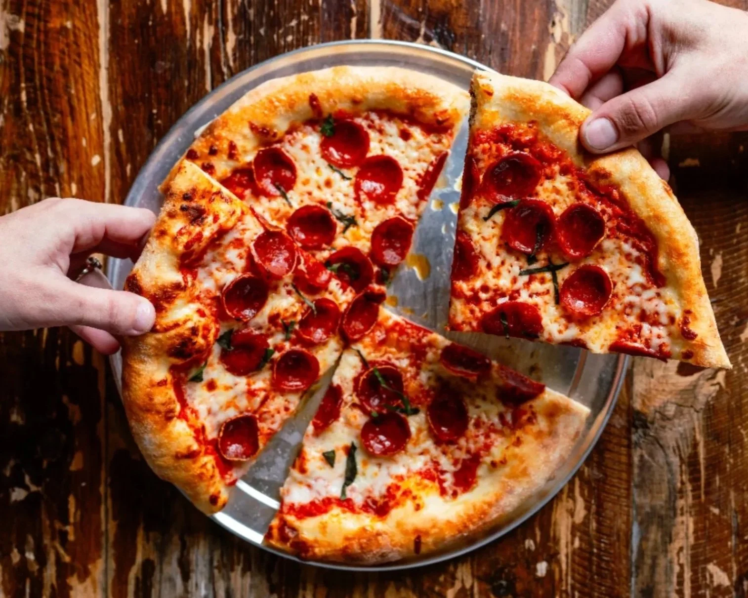 Two people sharing a pepperoni pizza from a metal pan on a rustic wooden table, with one person lifting a slice.