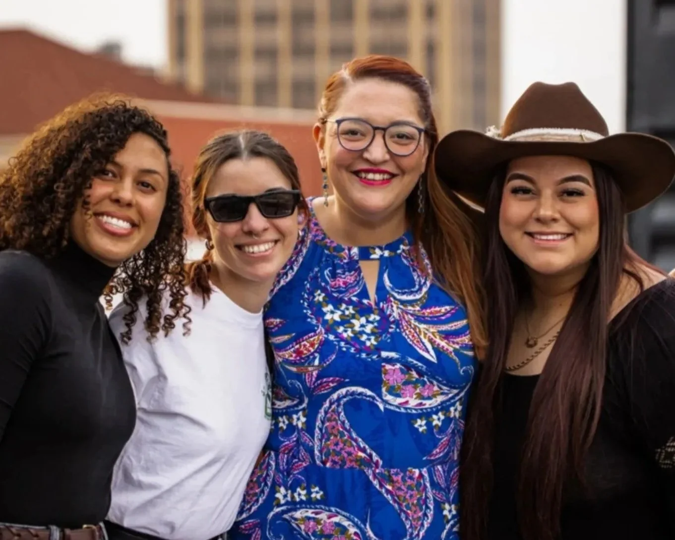 Four women standing together outdoors, smiling, with downtown Waco buildings in the background.