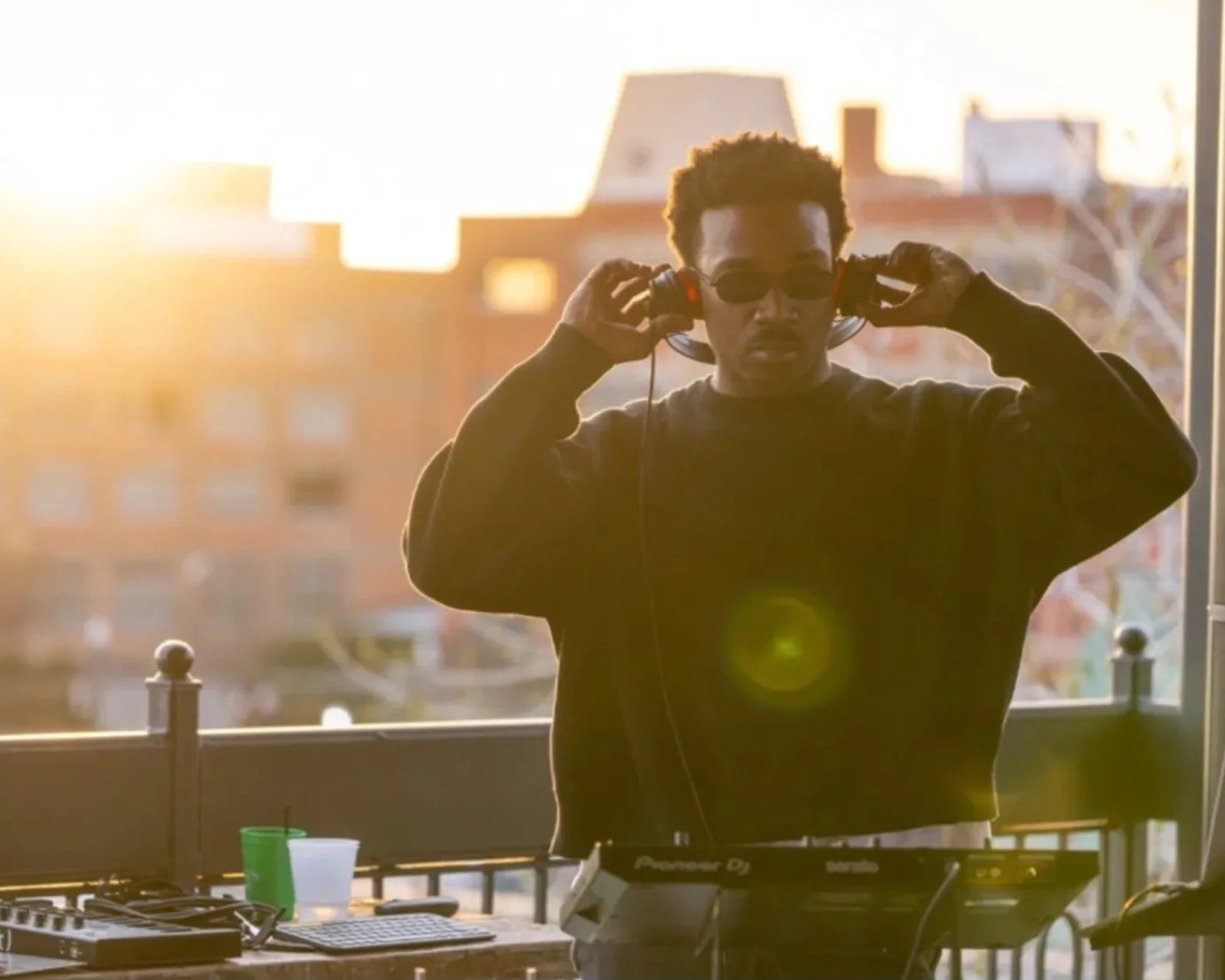 A DJ wearing sunglasses and headphones, standing outside at sunset with DJ equipment, a laptop, and drinks on the table.