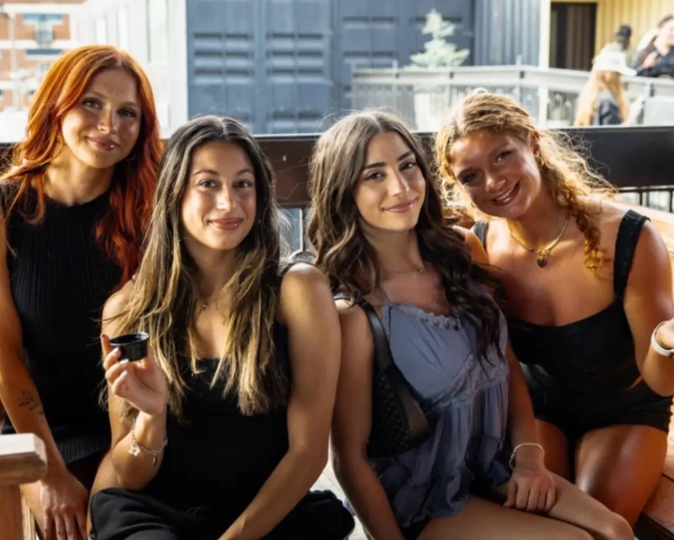 Four women sitting and standing together at an outdoor restaurant, smiling and holding small cups. Two women are sitting on a bench, and two are standing behind them. There are drinks and a straw on the table in front of them, with a blurred outdoor 