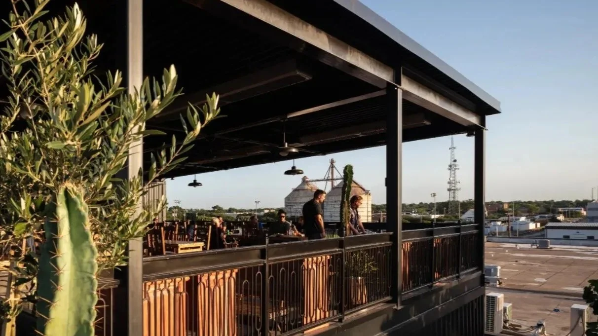 People on Lucky Buck's rooftop terrace in the late afternoon sun, with potted plants and cityscape in the background.