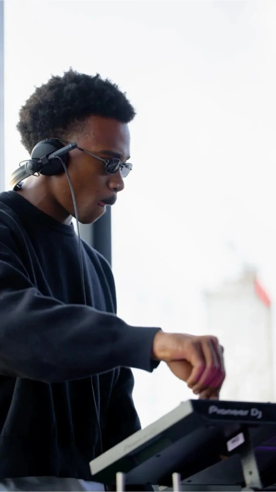 A young man with dark curly hair, wearing sunglasses and headphones, operating a DJ mixer or turntable.