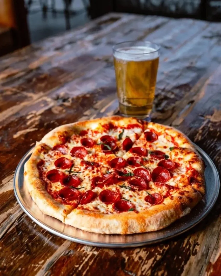 A pepperoni pizza on a metal tray with a glass of beer on a rustic wooden table.