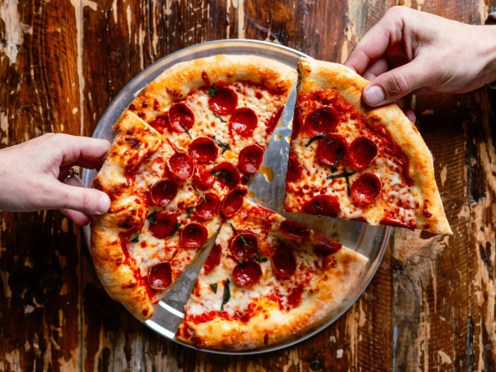 Two people sharing a pepperoni pizza from a glass platter on a rustic wooden table, with one person lifting a slice.