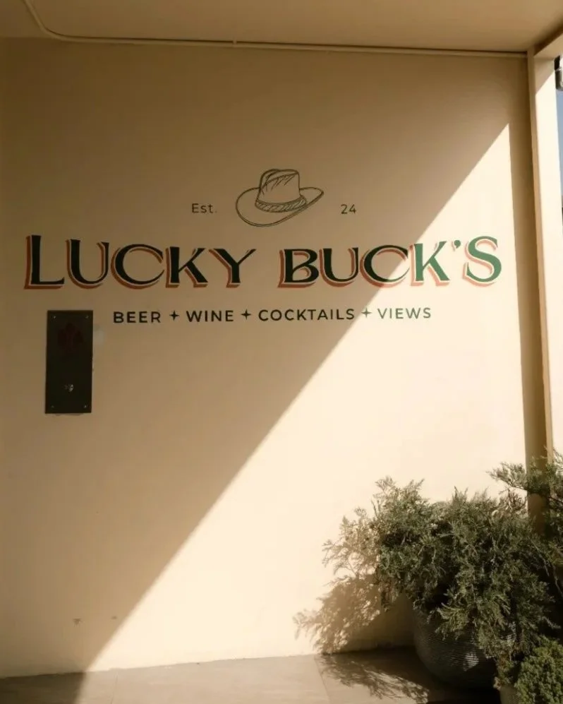A sign on a stucco wall of Lucky Bucks Rooftop logo with the words Beer Wine Cocktails and Views underneath with plants in the front of it.