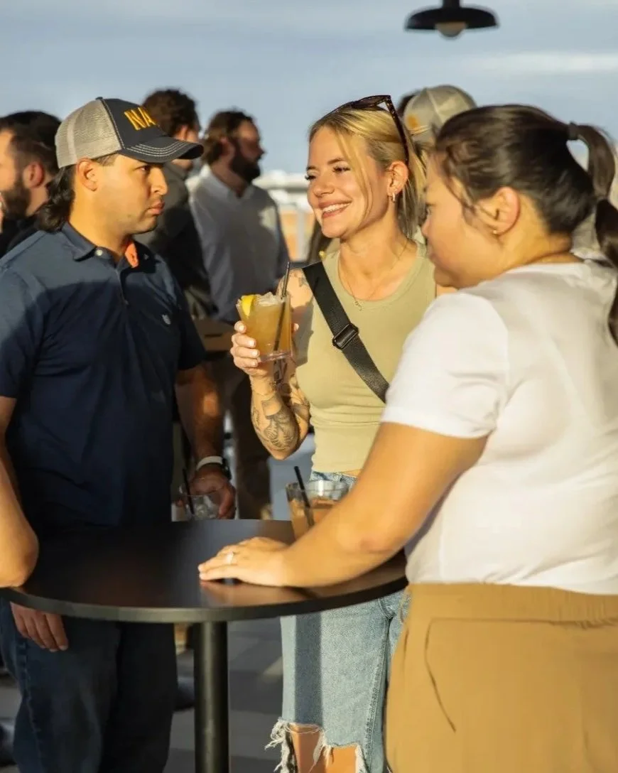 People socializing at an outdoor bar, with a woman smiling holding a drink, and two women and a man around a high table, in the evening with cityscape in the background.