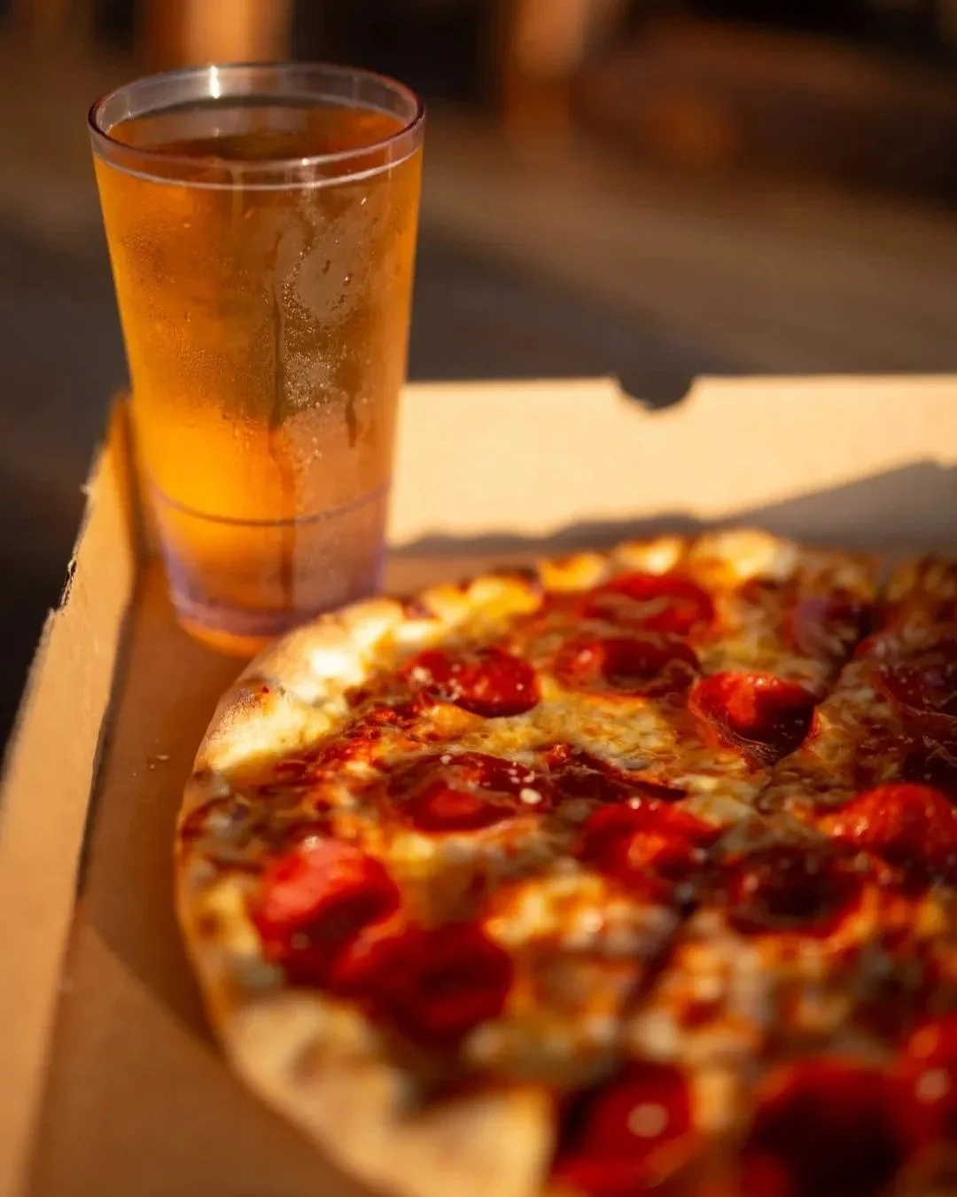 A slice of pepperoni pizza in a pizza box next to a glass of iced tea on a wooden table.