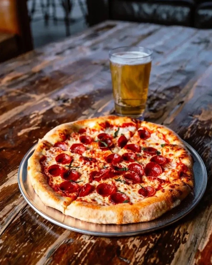 A pepperoni pizza on a metal tray with a glass of beer on a rustic wooden table.