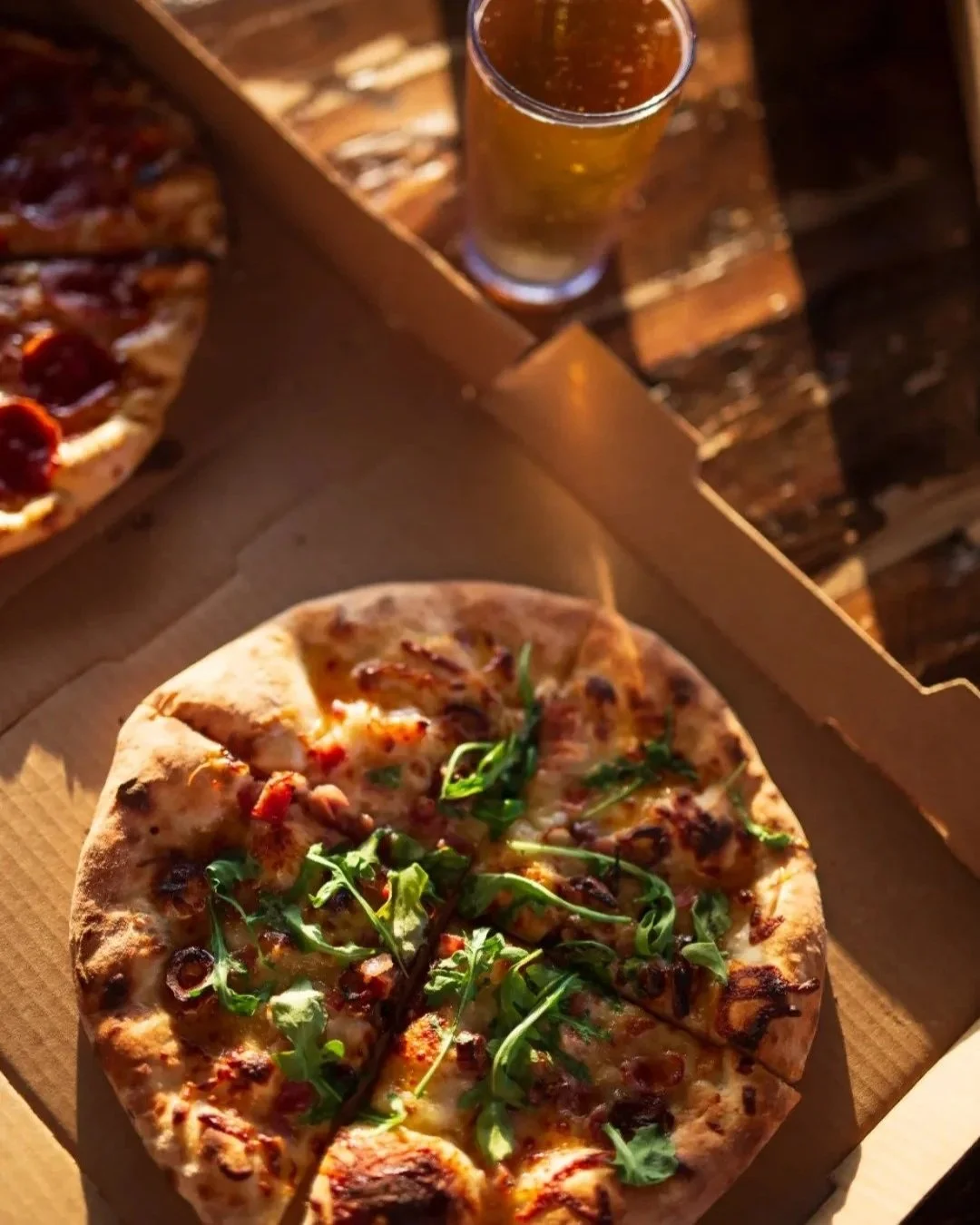 A pizza with arugula and bacon slices in a cardboard box, and a glass of beer on a wooden table.