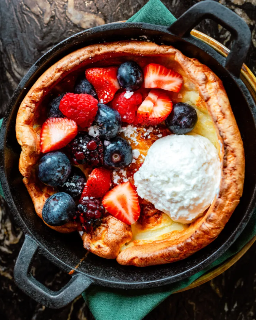 The Red Herring Dutch Baby Pancake served in a cast iron dish with fresh strawberries, blueberries and whipped cream. The pan is sitting on top of a green napkin.