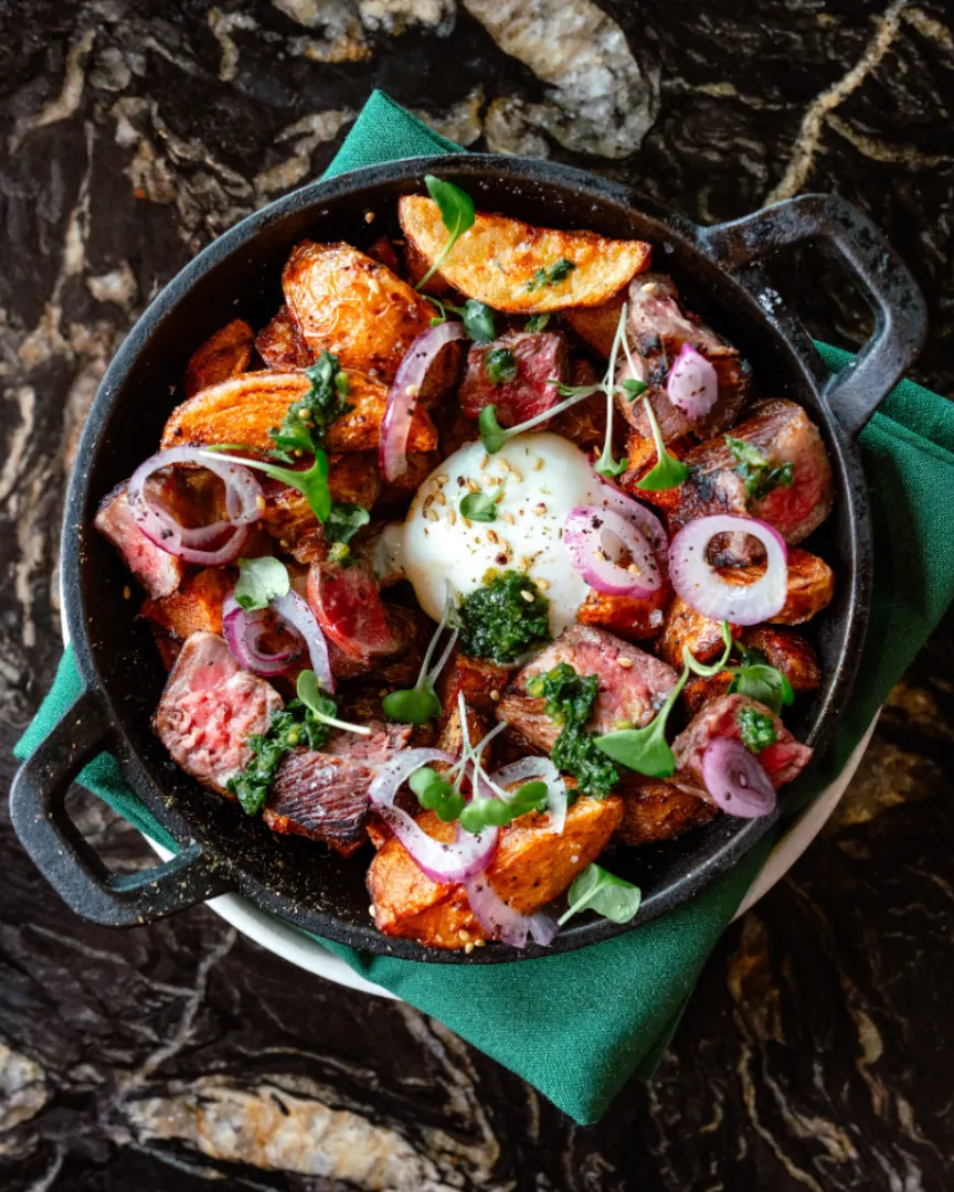 A brunch hash served in a cast iron pot on a green napkin. It features crispy potatoes and steak with a poached egg and red onions. There's green garnishes. Its on top of a black marble table.