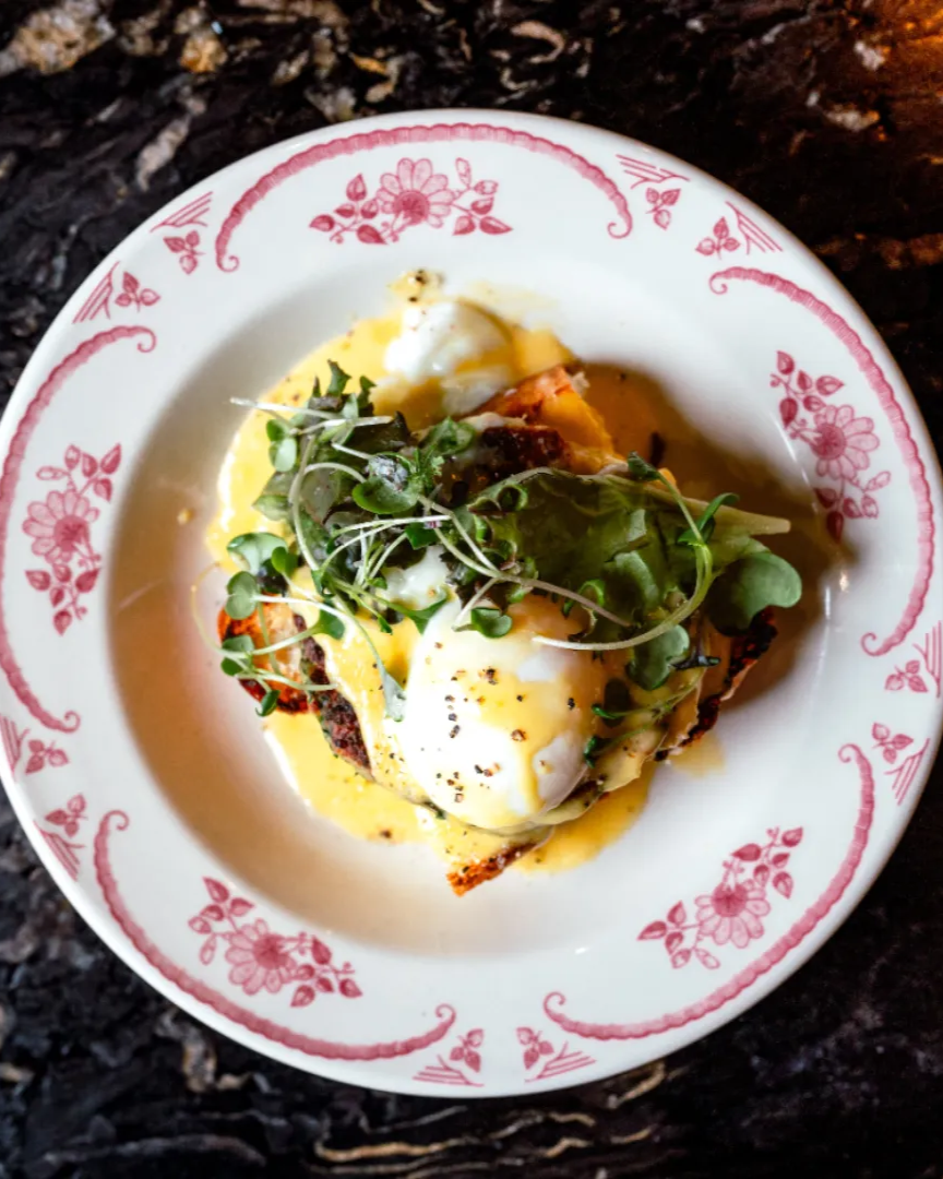 A brunch dish featuring a poached egg and hollandaise sauce with micro greens on top served on a white vintage plate with red scrolling elements and details, on top of a black marble table.