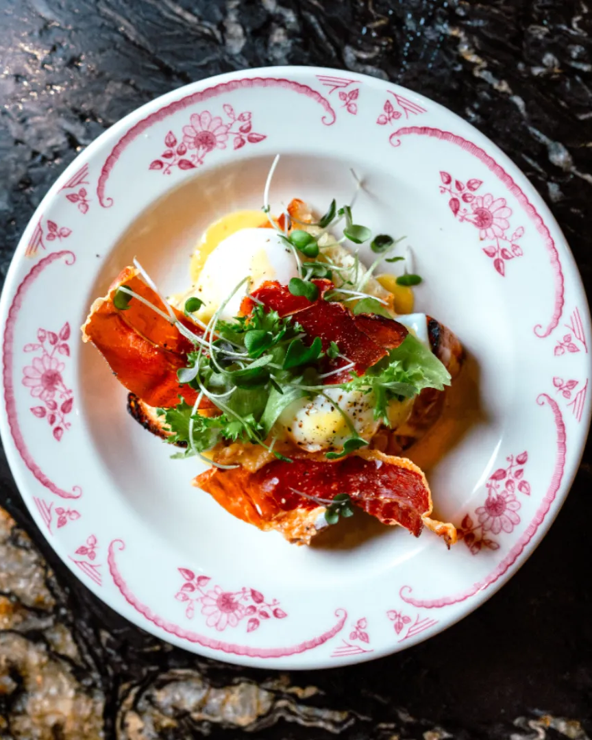 A delicious brunch plate on a vintage china plate with red flowers and scrolling lines, The plate features bacon and a poached egg with microgreens on top and cracked pepper.