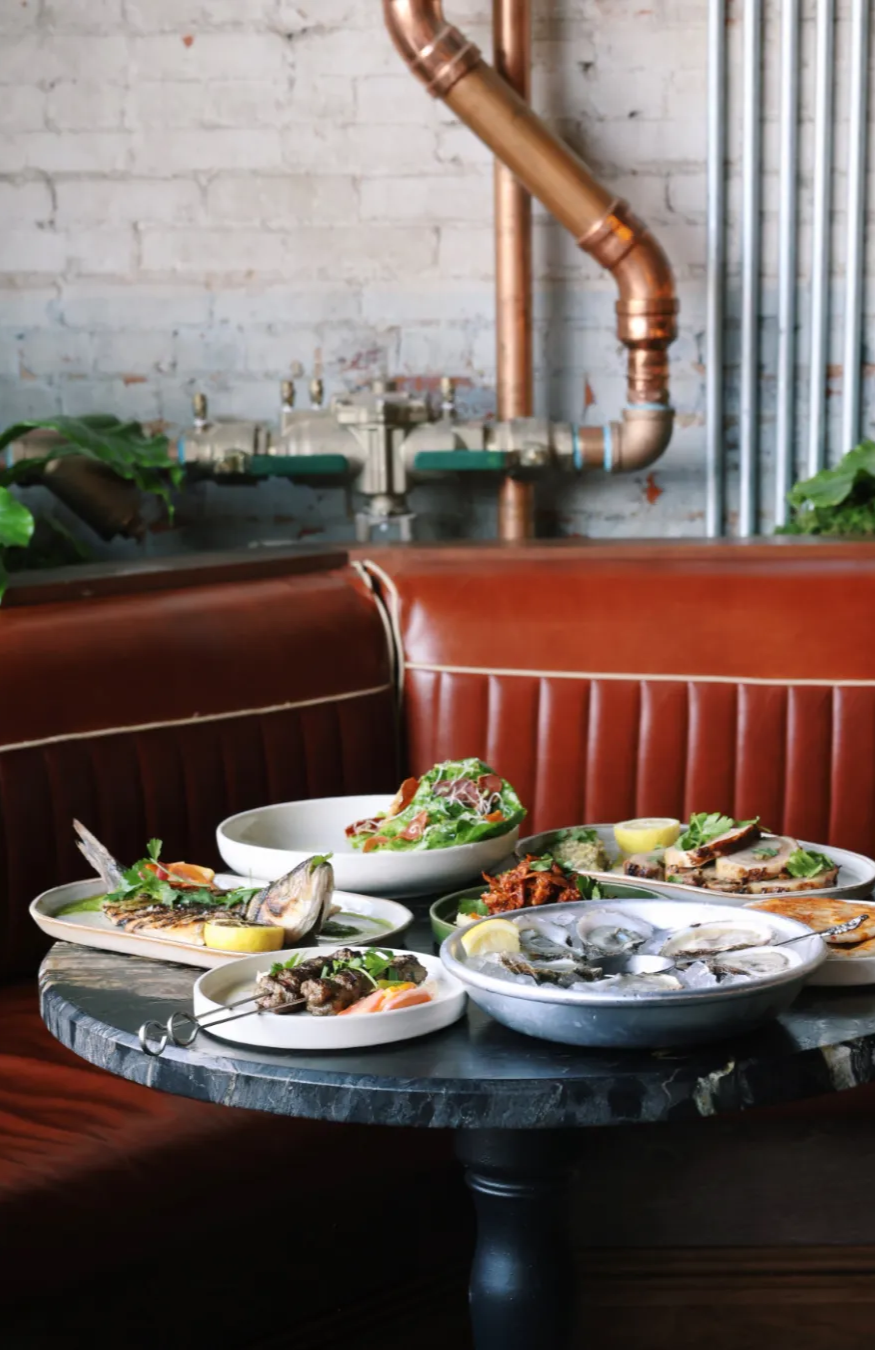 A view of the dishes on the table at Red Herring Restaurant surrounded by the leather booth seating and the industrial pipes and vintage brick at Red Herring's Cocktail bar. 