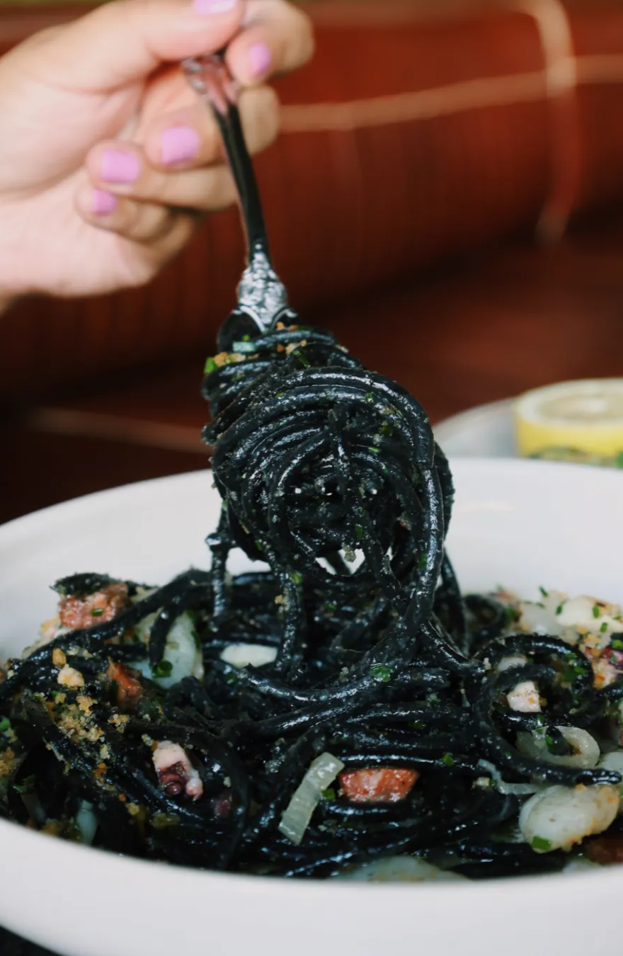 A hand wrapping up and pulling fresh squid ink pasta around a fork over the bowl. 
